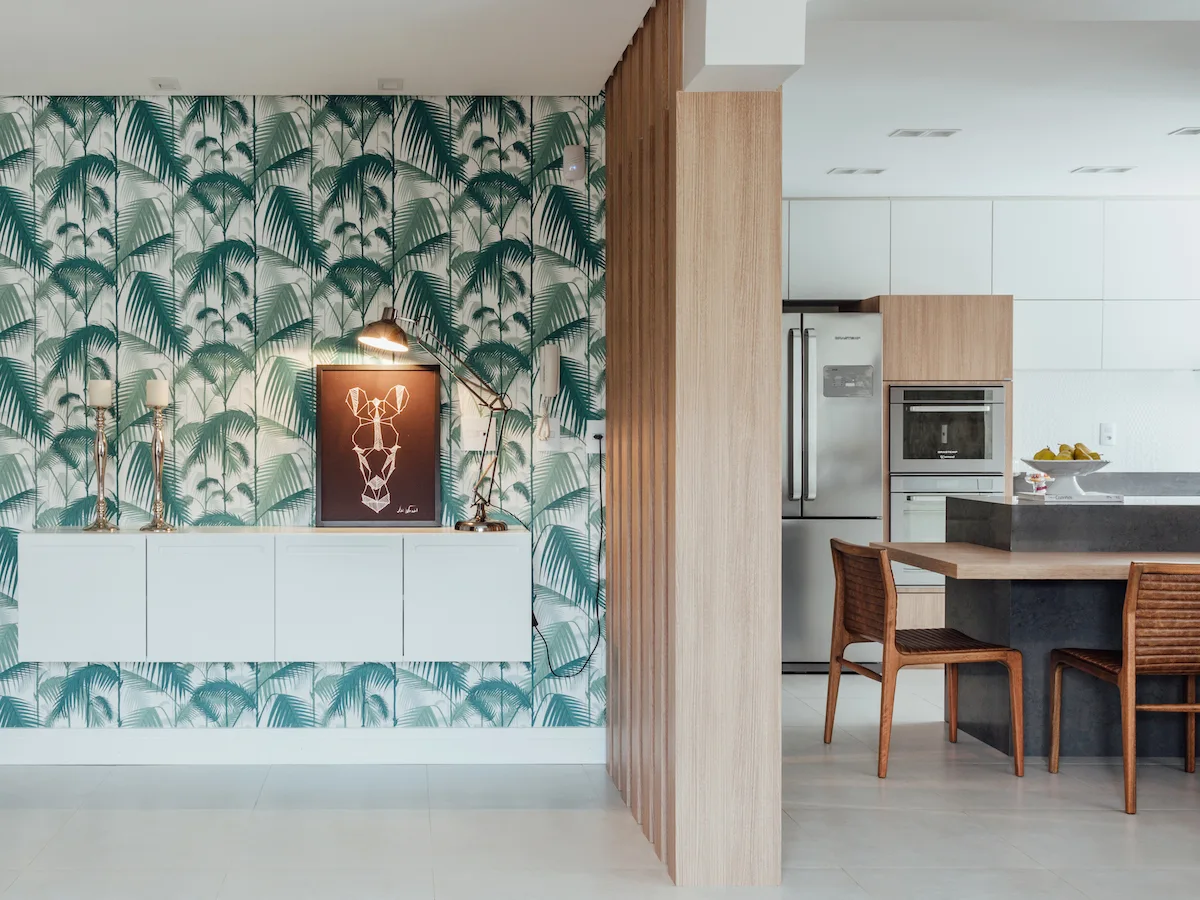 A floating white console sits against green palm leaf wallpaper next to a slatted wood partition that opens into a modern kitchen with a grey island.