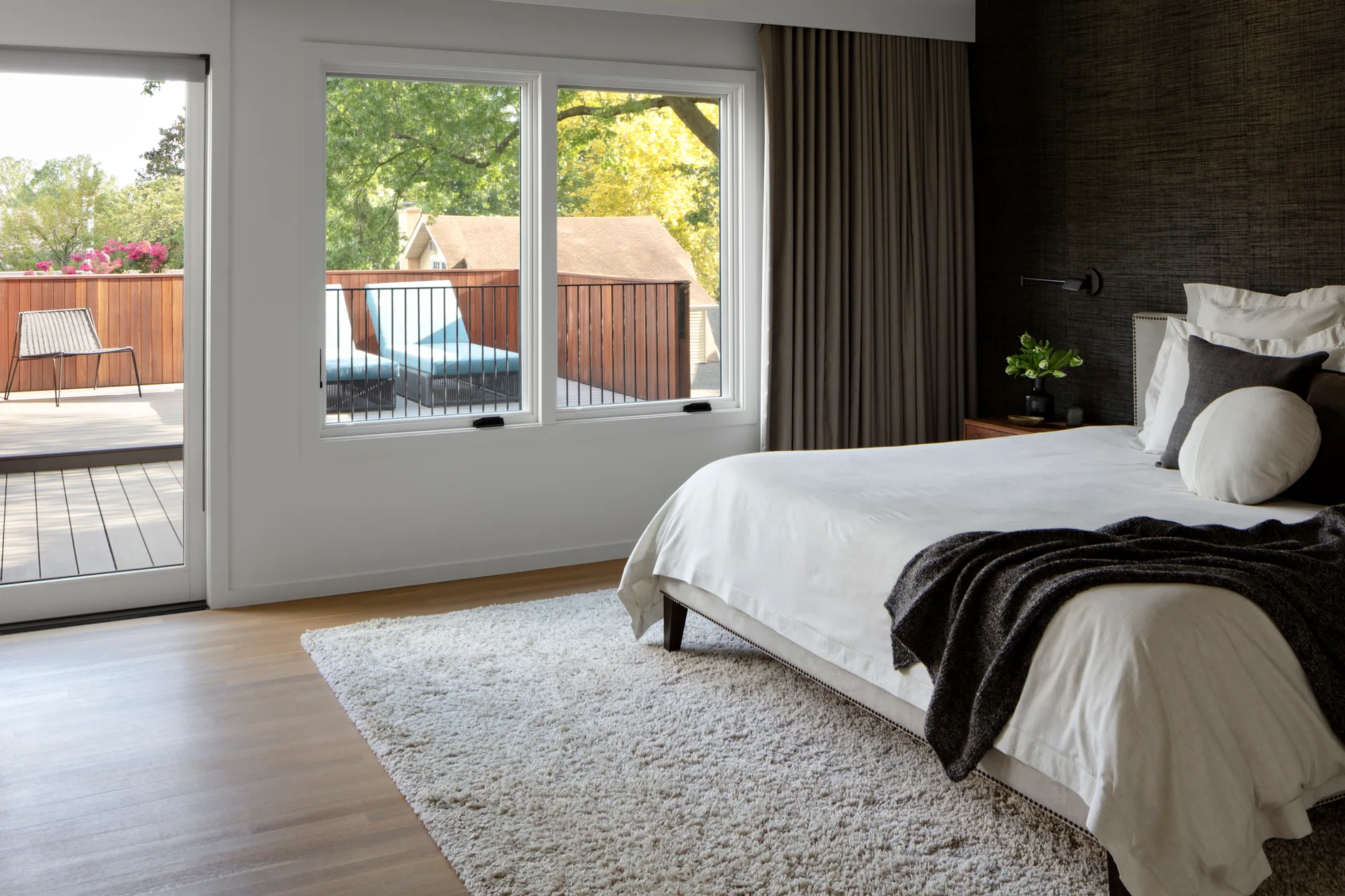 A modern bedroom features a white upholstered bed against a dark textured wall next to large windows overlooking a wooden balcony.