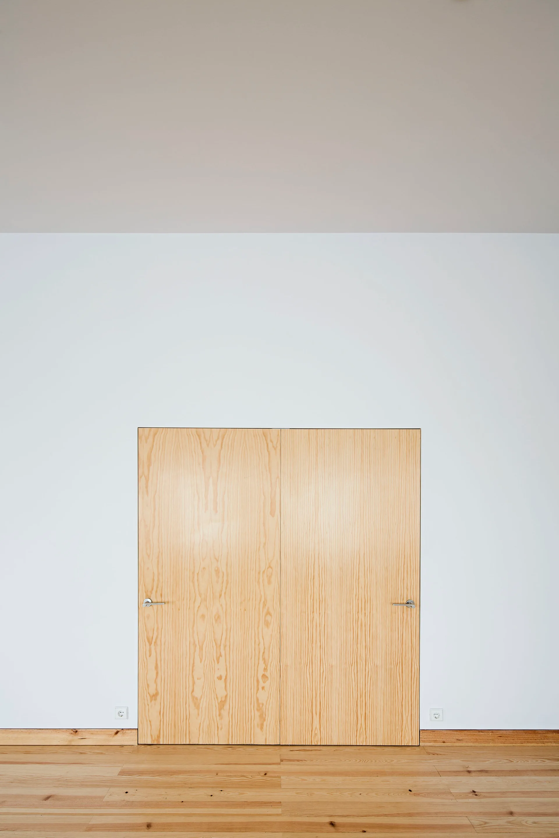 A minimalist white room showing a flush light-toned wooden double door with silver handles and matching wood floors.