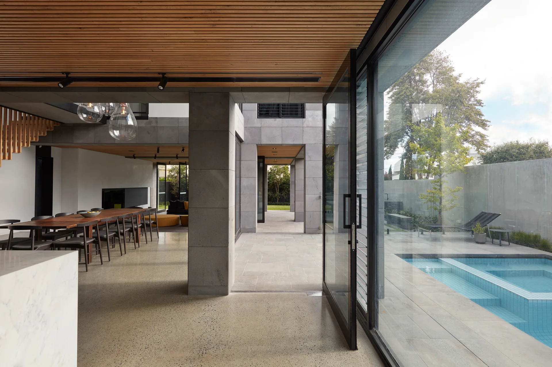 An open indoor dining room with a wood slatted ceiling and concrete floors opens up to a paved outdoor terrace with a swimming pool.