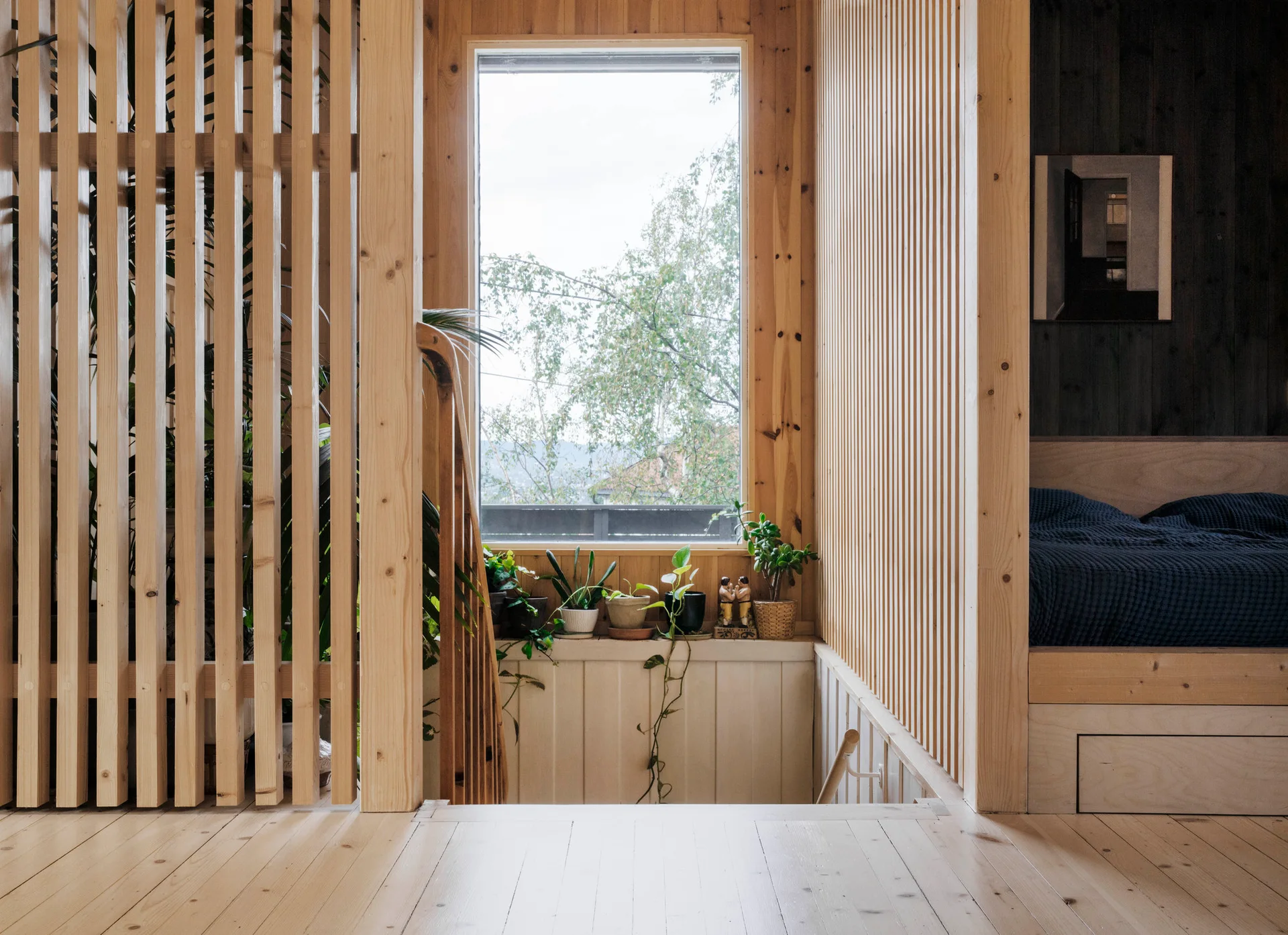 A minimalist light wood bedroom featuring vertical slatted wood partitions, a built-in alcove bed with blue sheets, and plants on a windowsill.