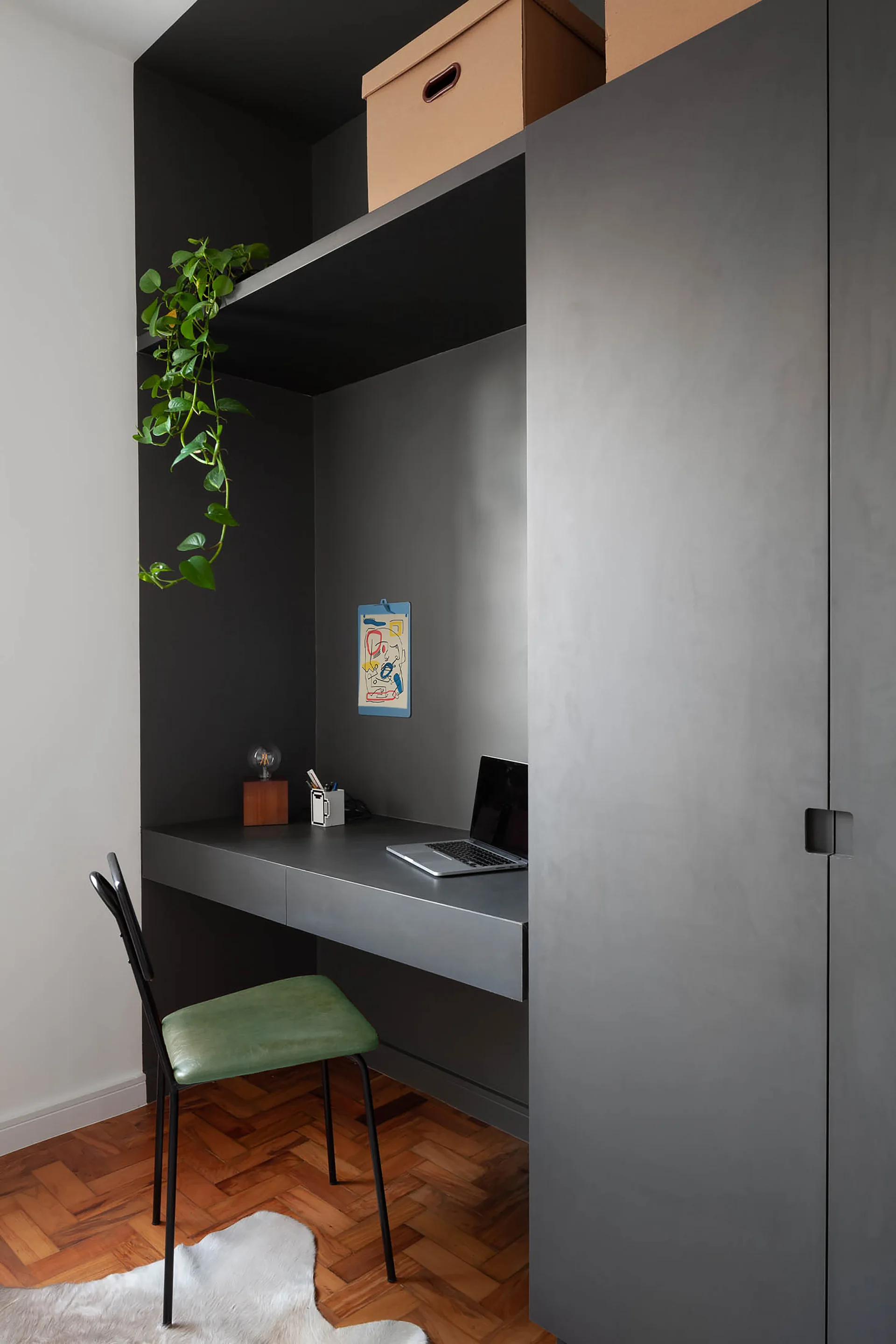 A matte charcoal built-in desk and wardrobe niche features a green chair, herringbone wood floor, and a laptop on the desk surface.