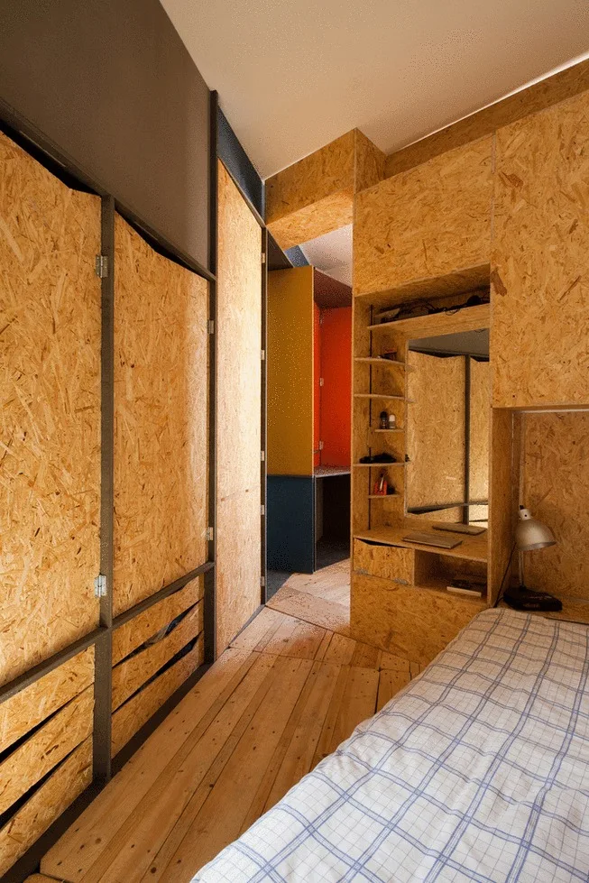 A compact bedroom featuring custom built-in cabinetry made of oriented strand board, a bed with checkered bedding, and a wooden floor.