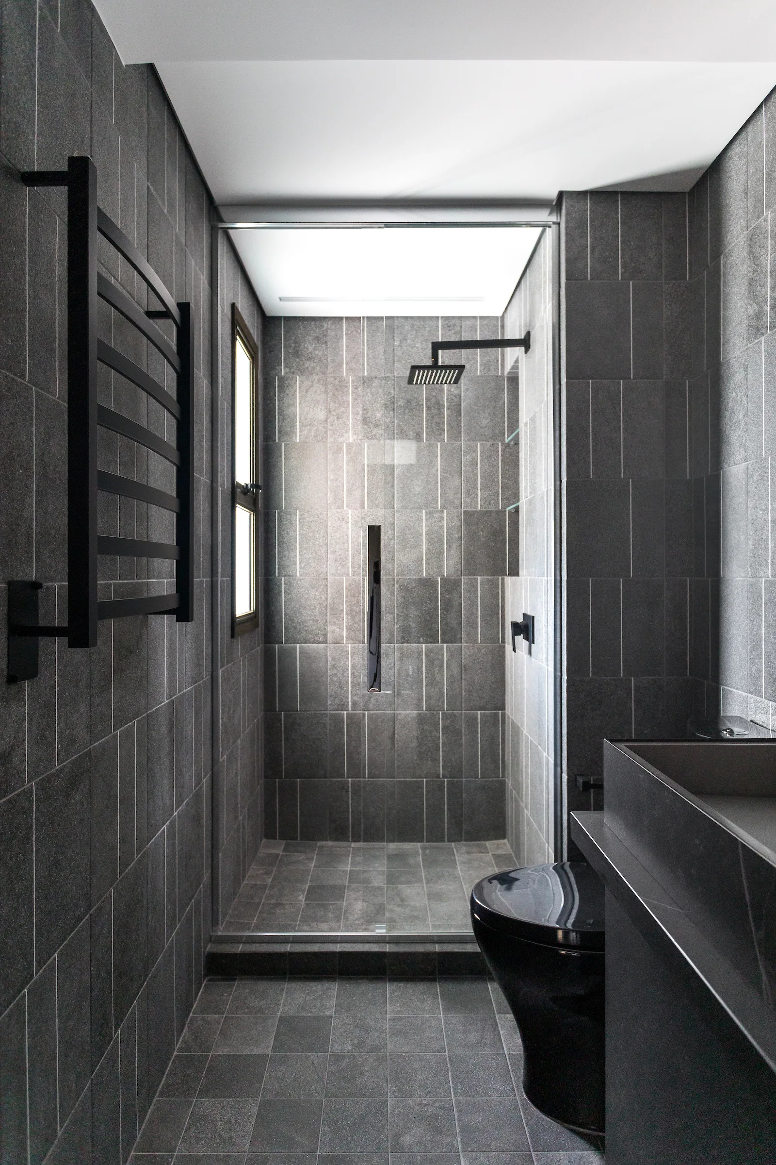 A narrow modern bathroom lined in dark grey rectangular stone tiles features a walk-in shower, a black toilet, and a matte black ladder-style towel warmer.