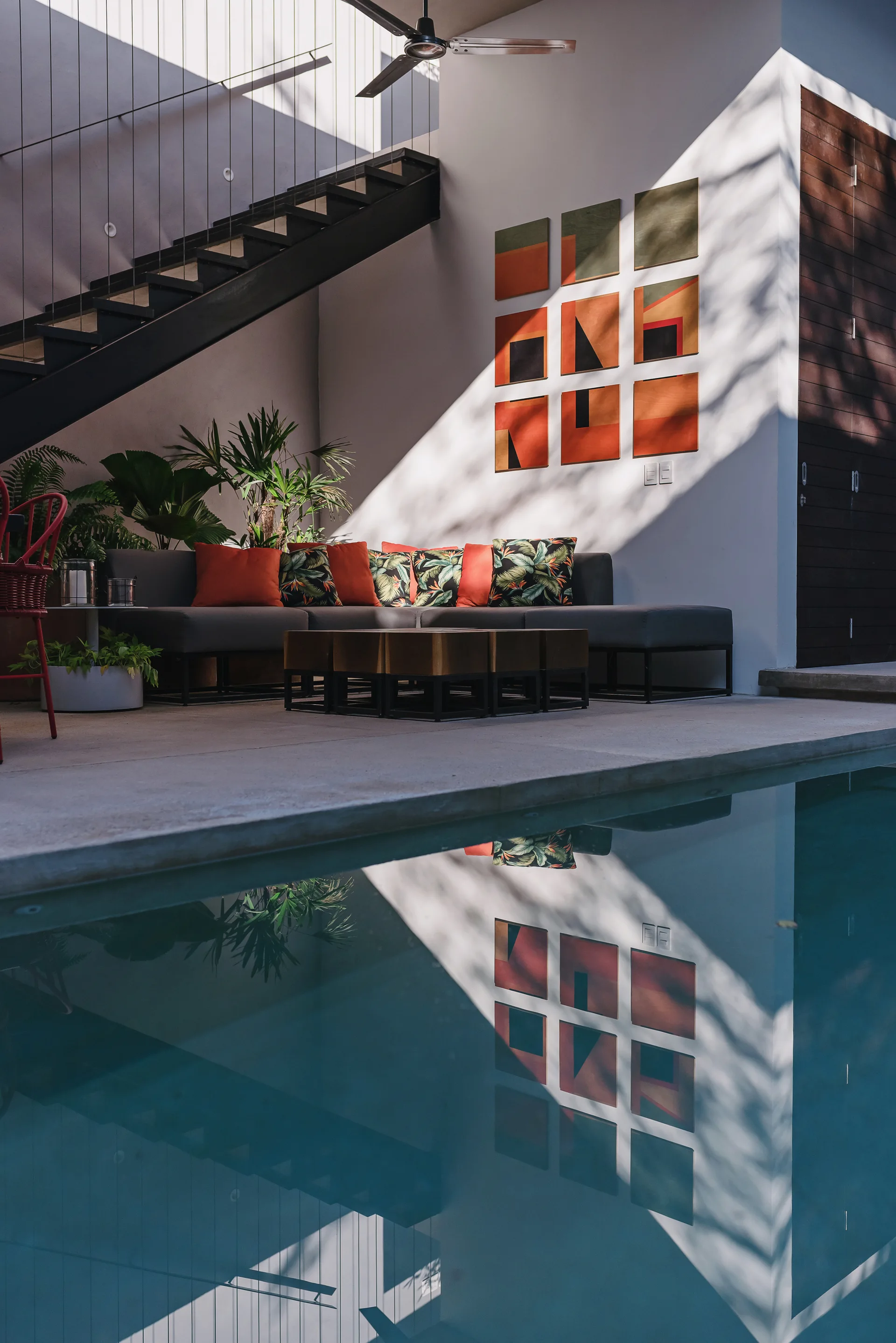 A poolside lounge area features a gray sectional sofa, a grid of colorful wall art, and a reflective swimming pool.