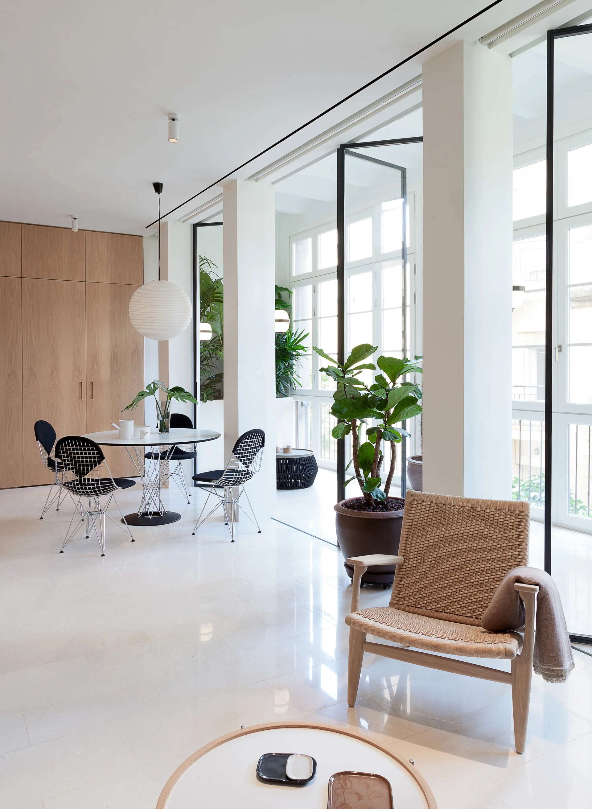 A bright minimalist living area features a woven wood armchair in the foreground, wire-frame dining chairs in the back, and tall glass doors.