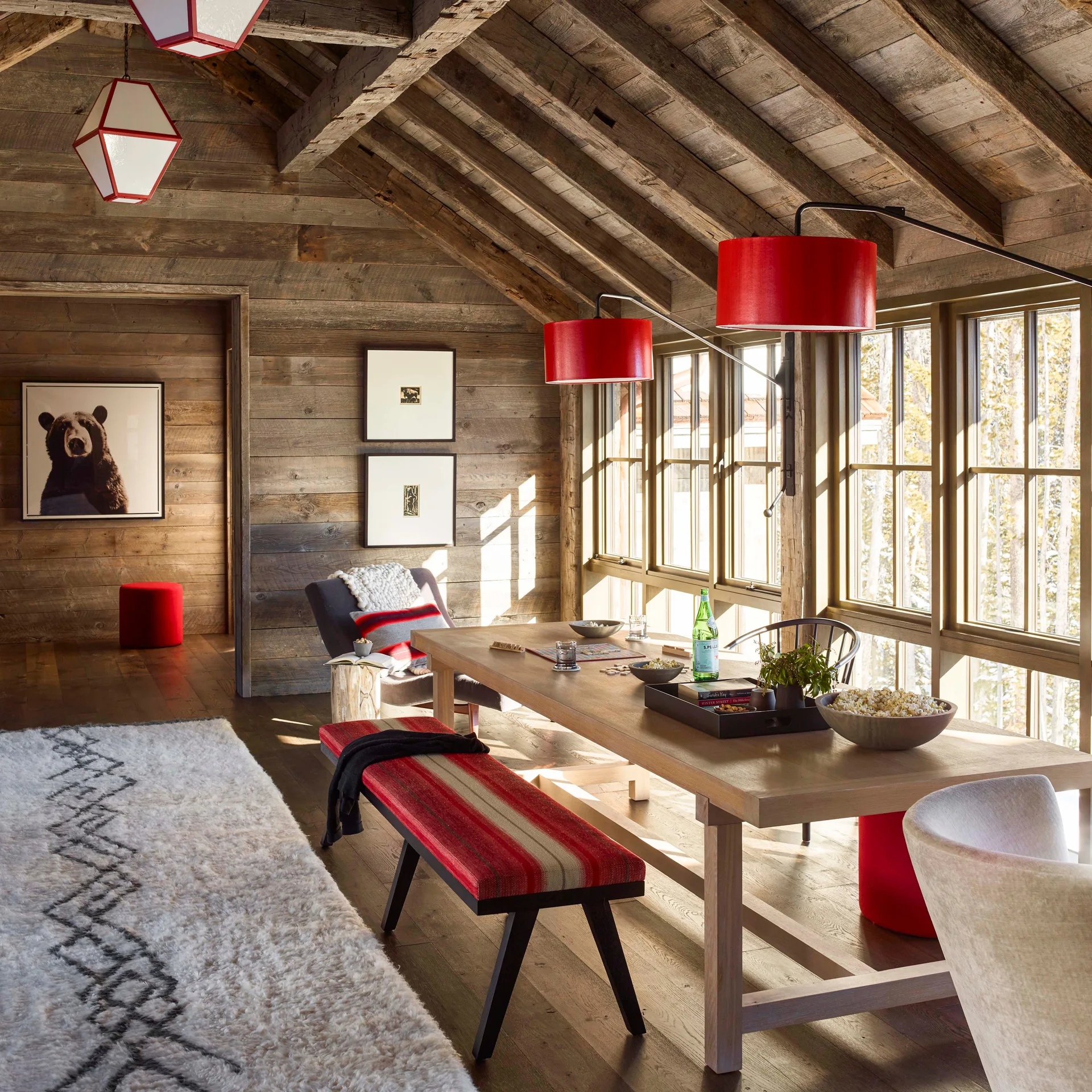 A rustic dining room featuring a long wooden table, a striped bench, wood-paneled walls, and large windows with red pendant lights hanging above.