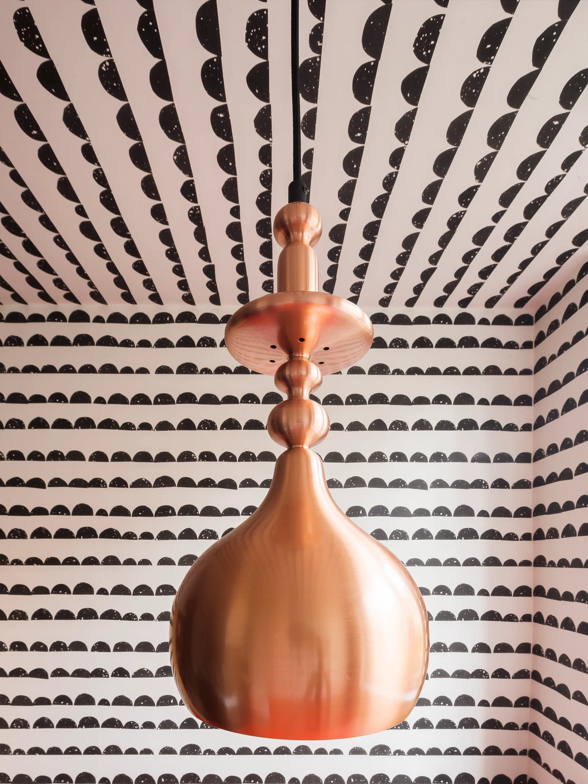 A sculptural copper pendant light hangs in front of walls and a ceiling covered in a repeating black-and-white semicircle patterned wallpaper.