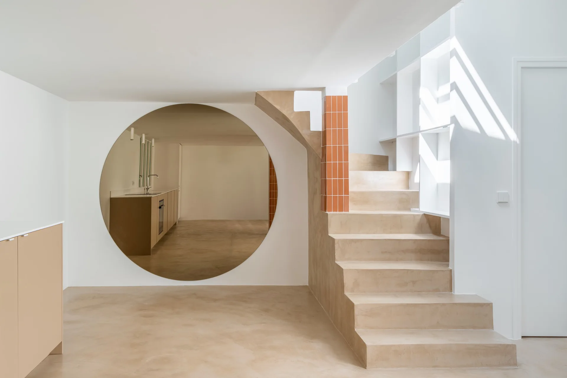 A minimalist interior featuring a smooth beige concrete staircase, a large circular wall mirror, and an orange-tiled architectural column.