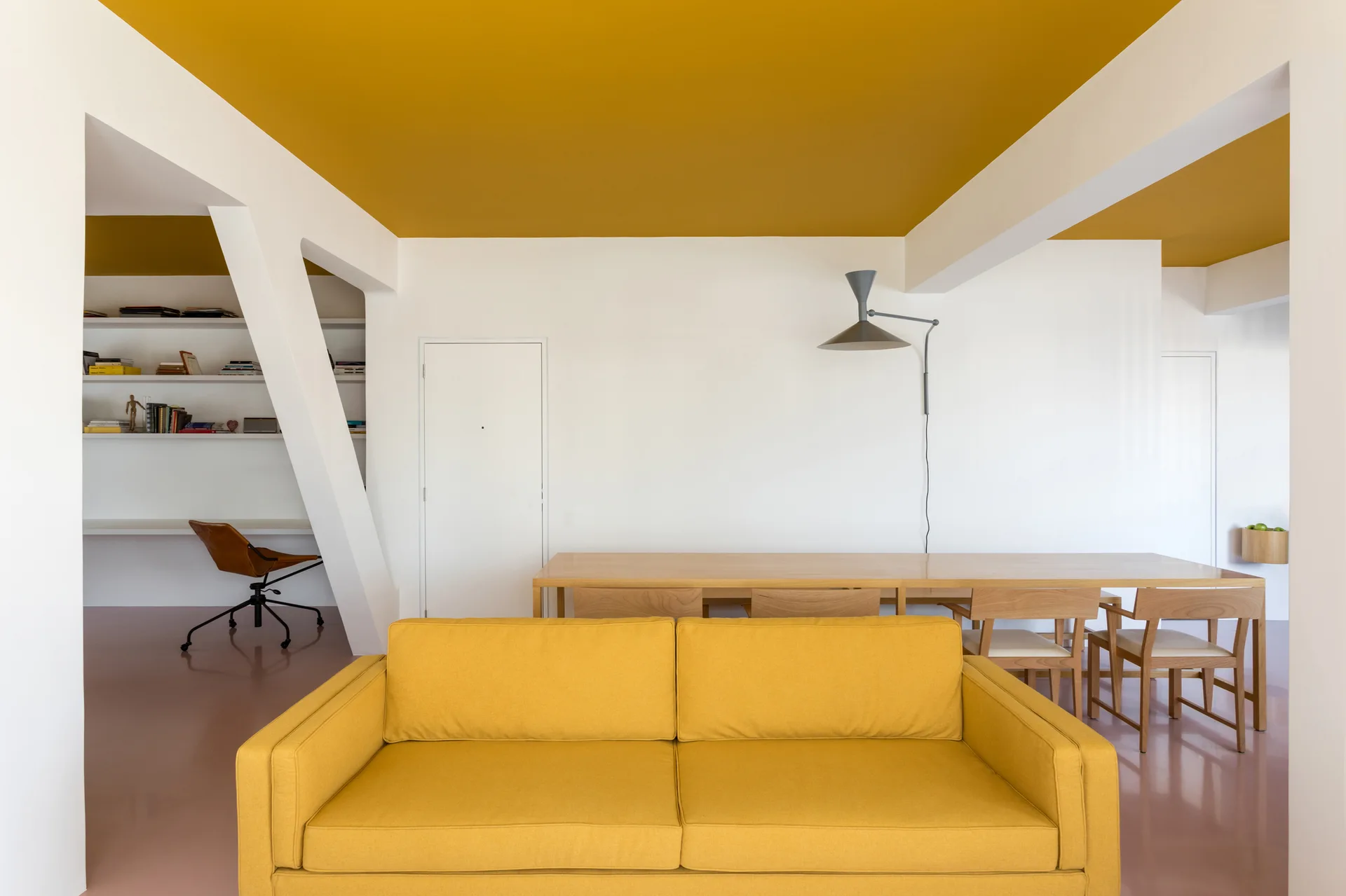 A modern living room features a bright yellow sofa, a wooden dining table, white walls, a diagonal column, and a mustard yellow ceiling.