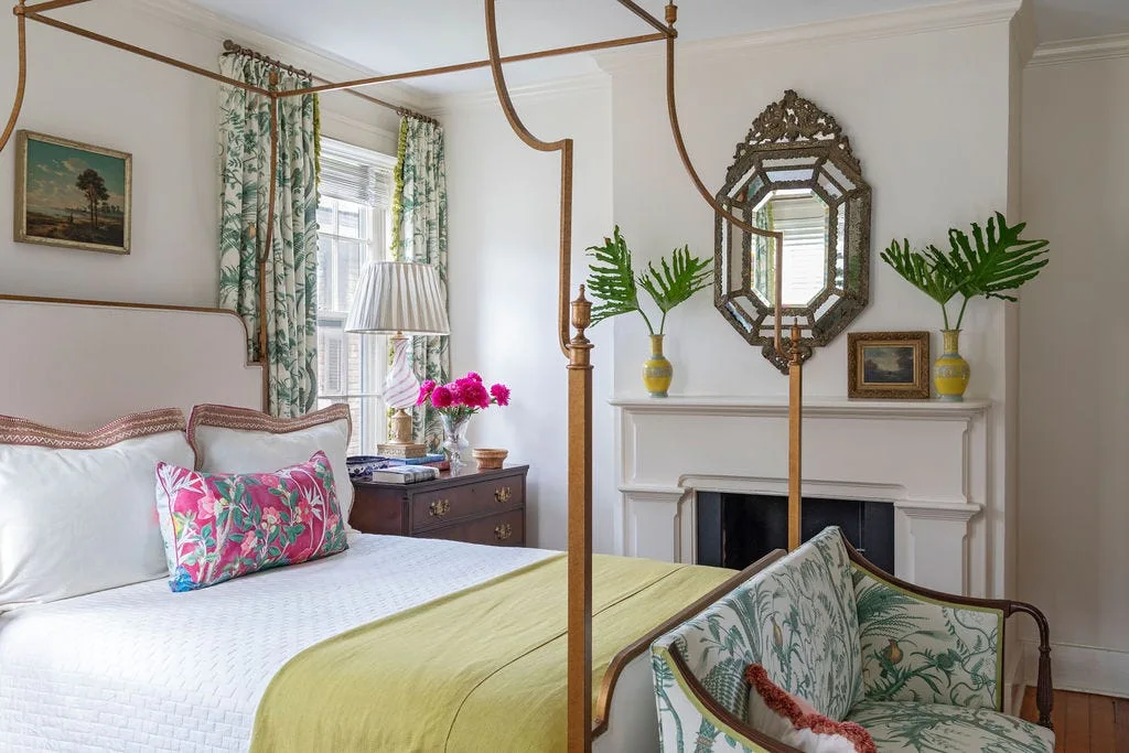 A classic bedroom featuring a gold four-poster bed, floral print curtains, a dark wood nightstand, and an ornate mirror hanging above a white fireplace.