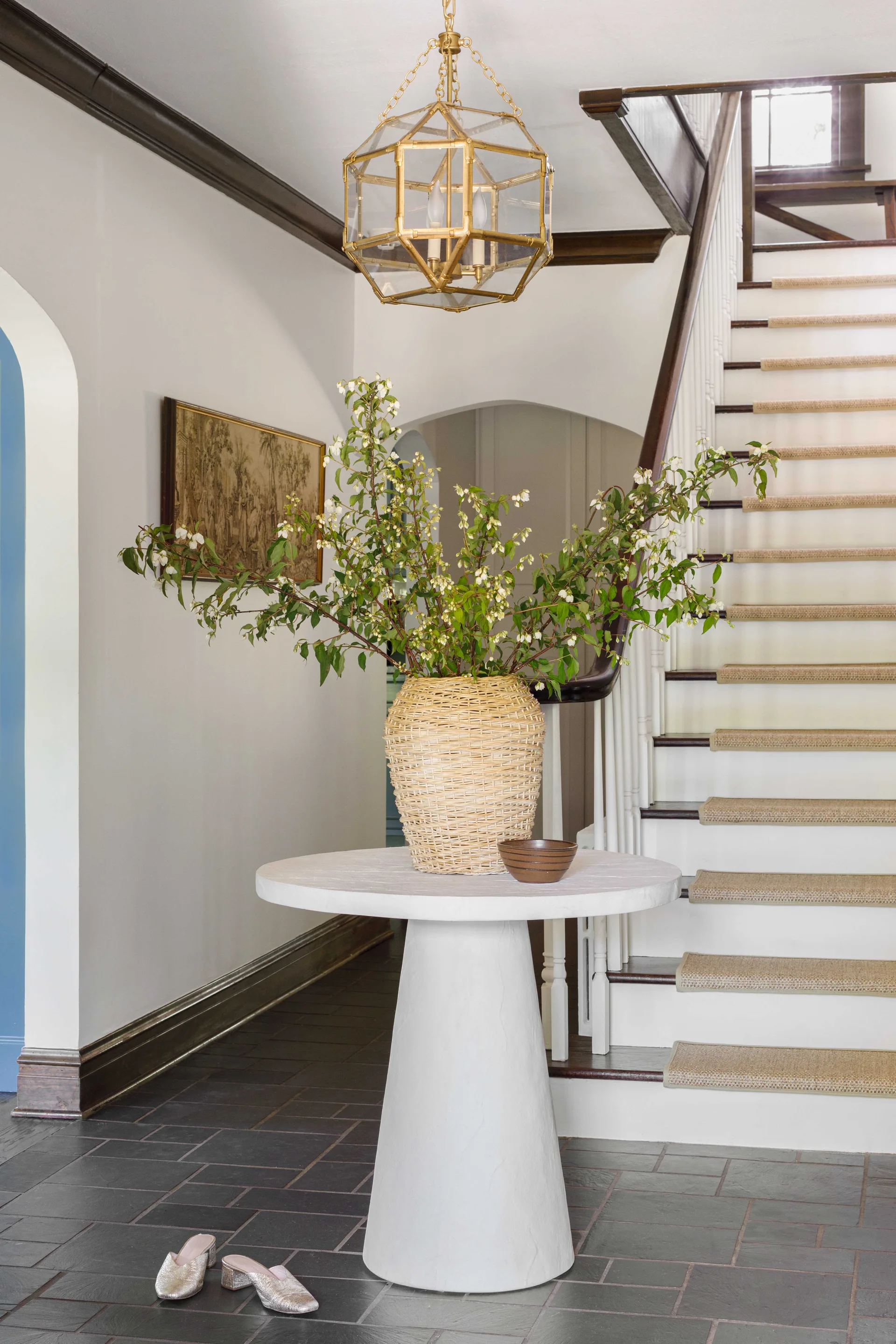 A white pedestal table holding a woven wicker vase with tall branches stands in an entryway with dark slate flooring and a staircase.