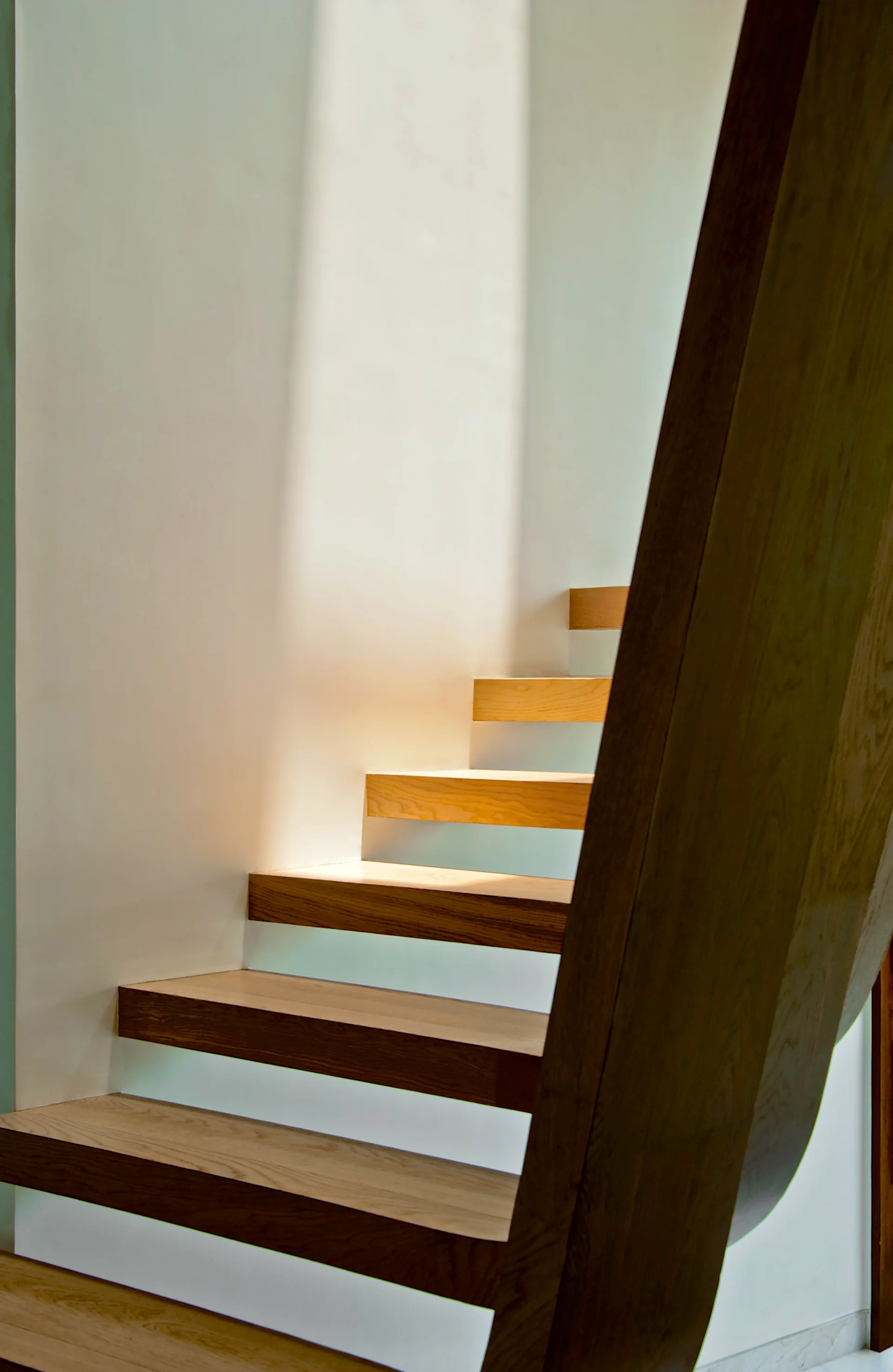 A close-up view of a modern floating staircase featuring thick wood treads alongside a smooth white wall under soft natural light.