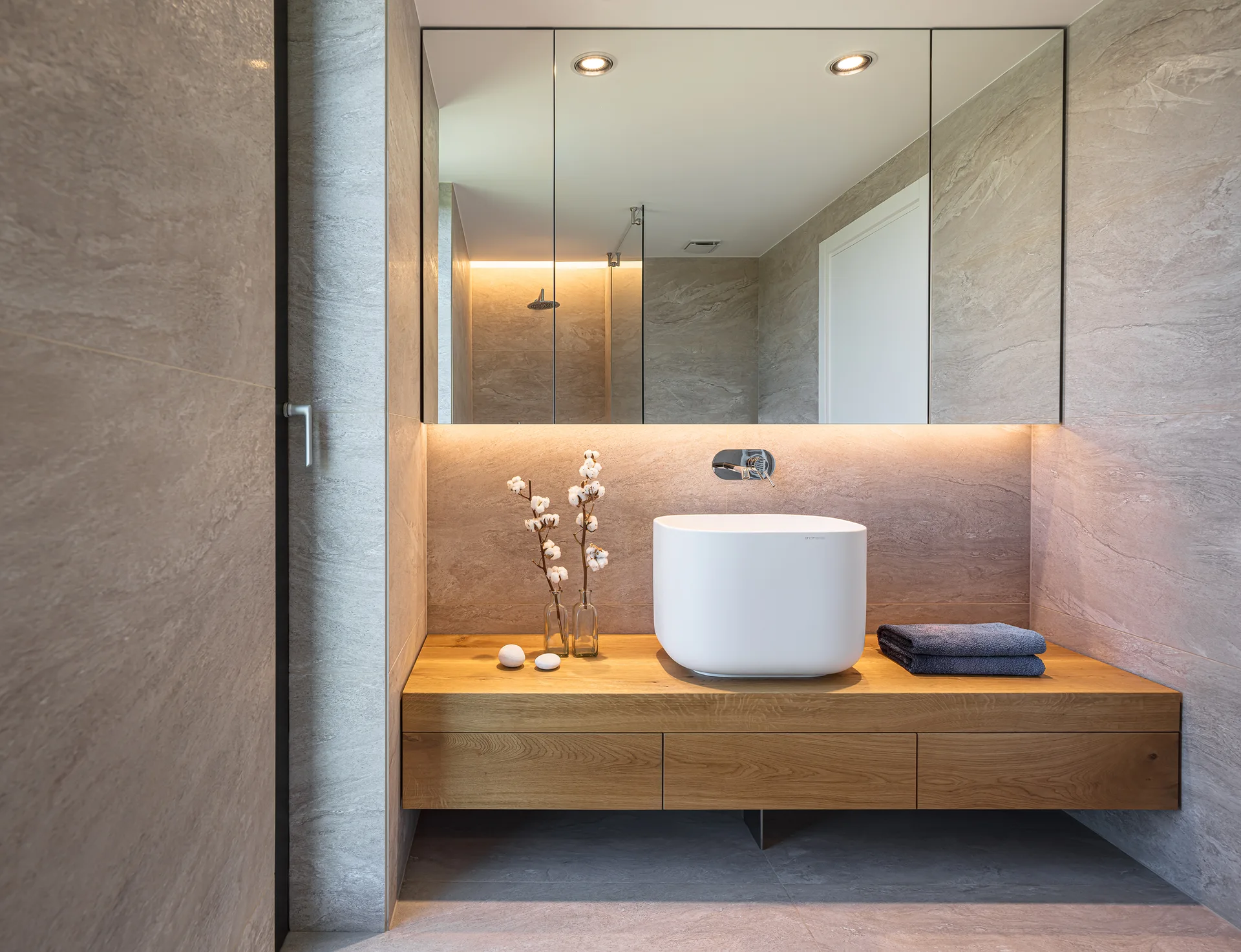 A modern bathroom features a floating wooden vanity with a white vessel sink, a large backlit mirror, and grey stone-tiled walls.