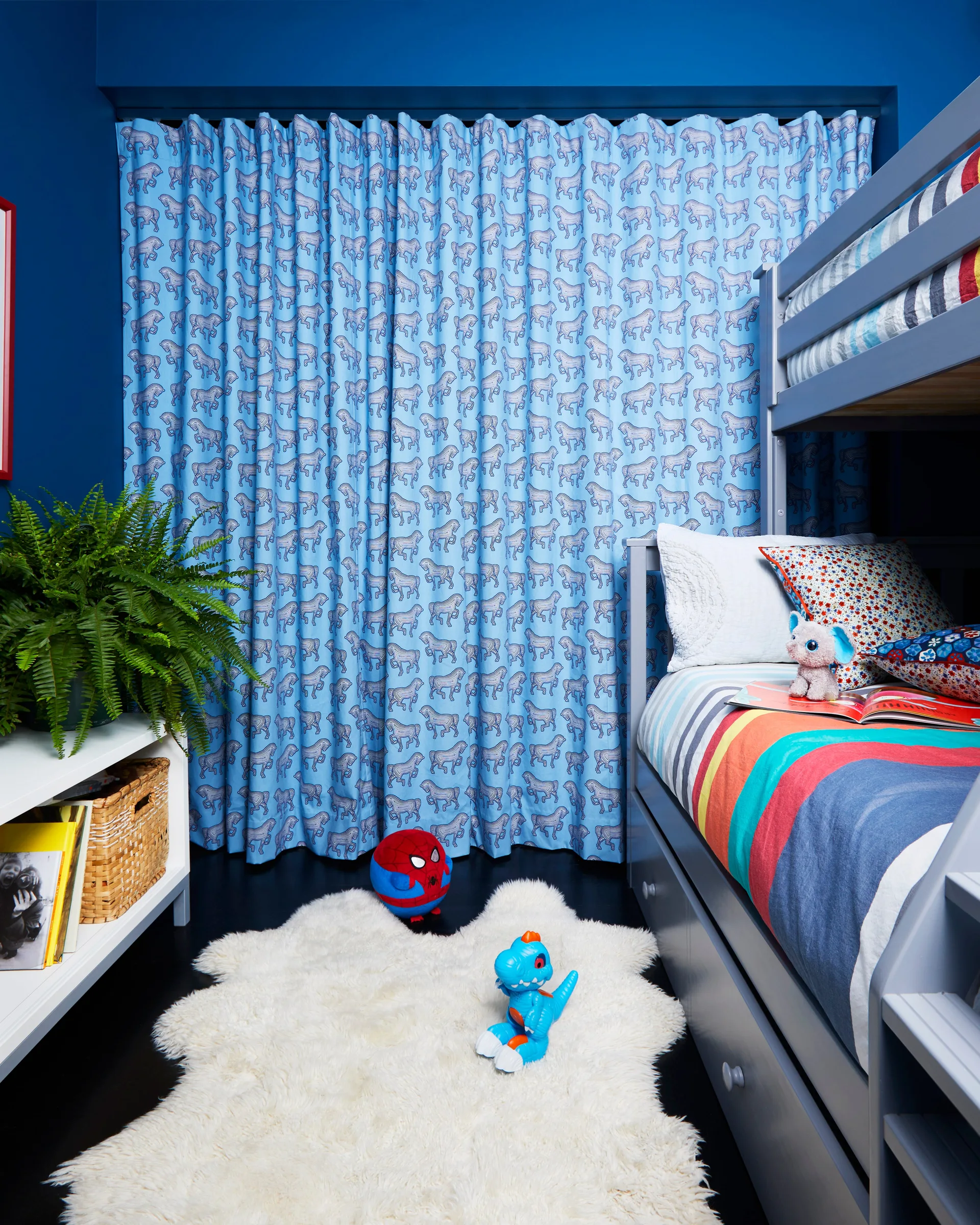 A children's bedroom with deep blue walls, a grey wooden bunk bed, blue animal-patterned curtains, and a white sheepskin rug.