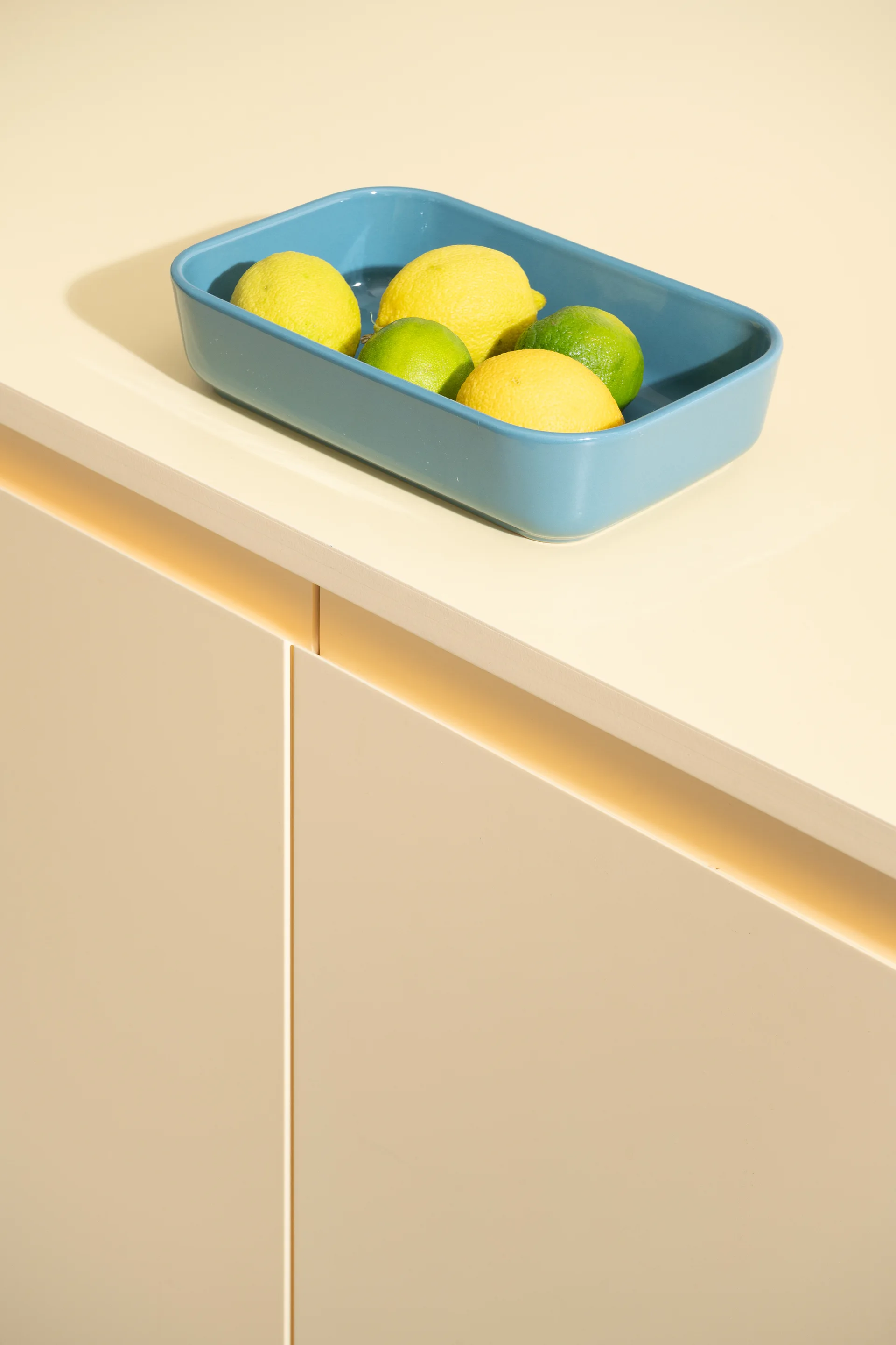 A blue rectangular ceramic dish filled with lemons and limes rests on a smooth cream-colored kitchen countertop above flat-front cabinets.