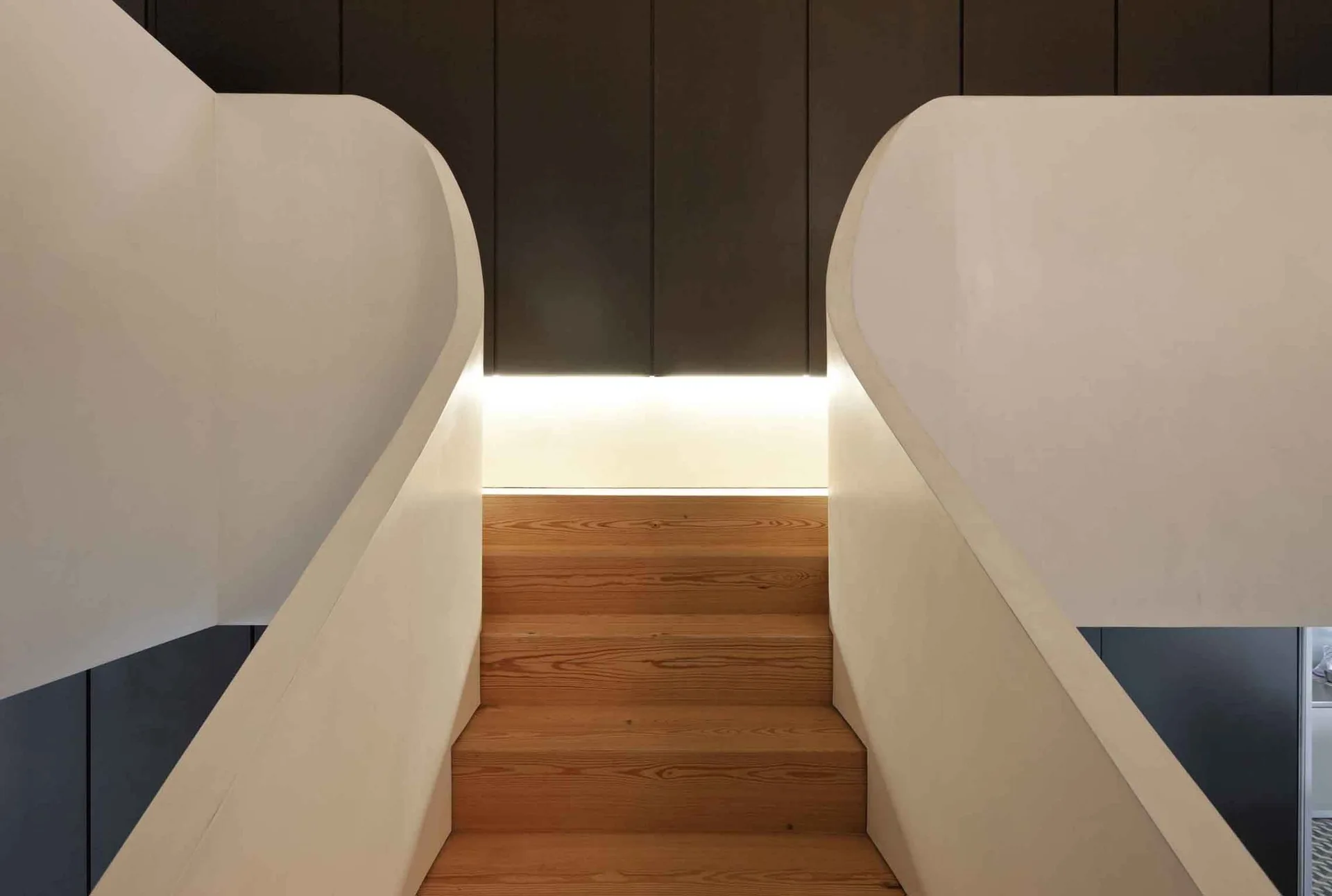 A modern staircase features warm wood treads between two sweeping, curved white walls under soft cove lighting against dark vertical paneling.