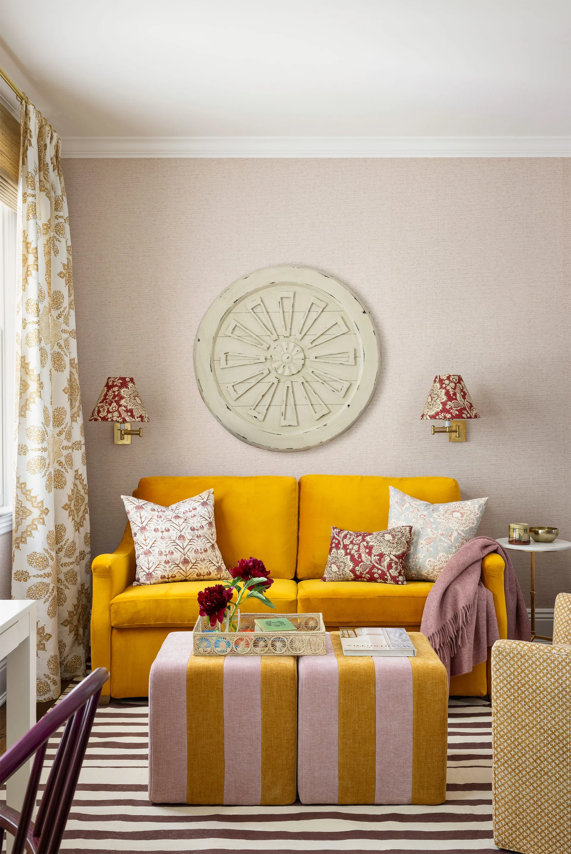 An eclectic living room features a bright yellow velvet sofa, two pink and yellow striped ottomans, and a round wooden medallion on the wall.