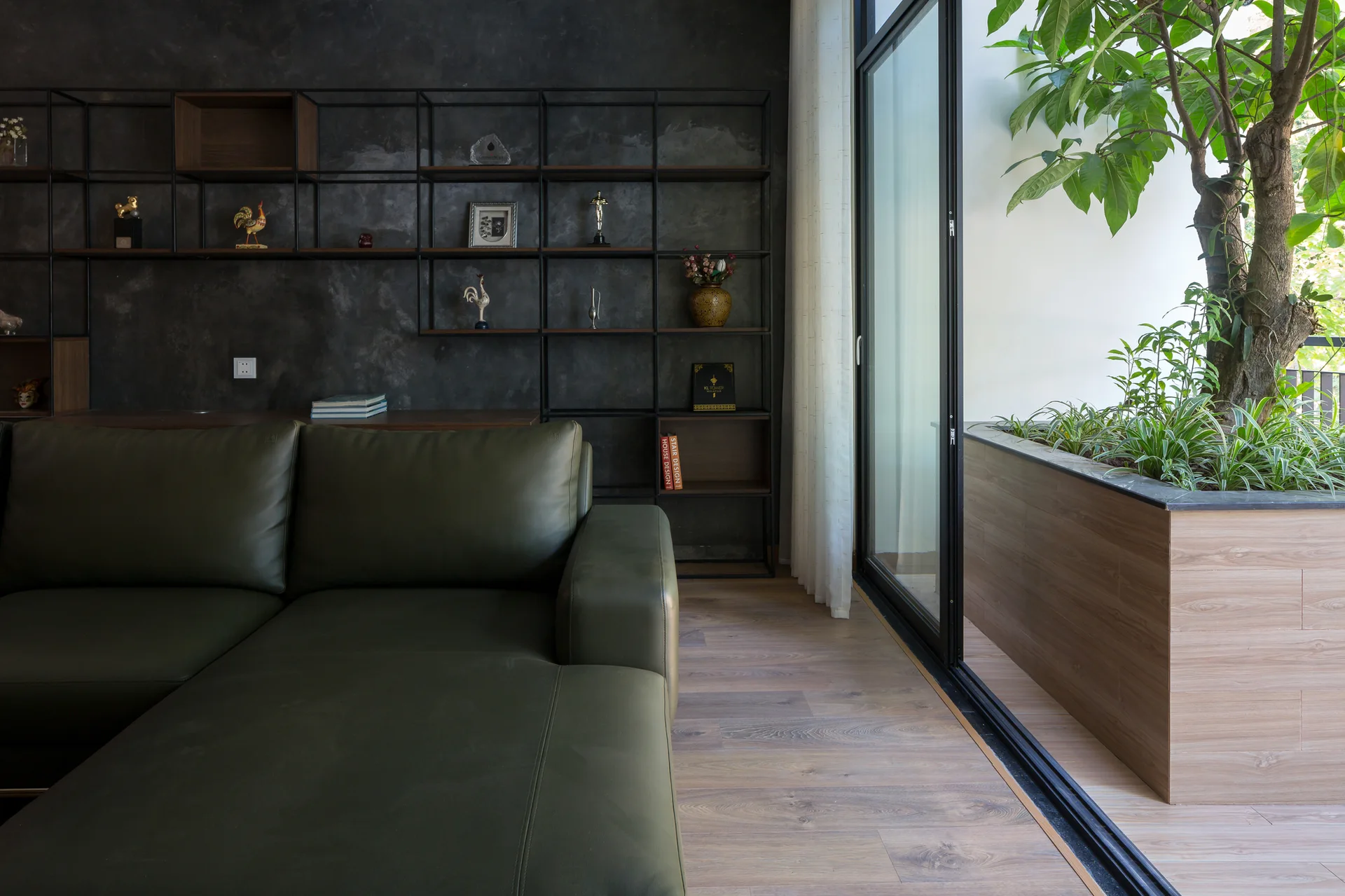 A green leather sectional sofa sits on light wood flooring in front of a dark textured accent wall with black metal shelving next to a sliding glass door.