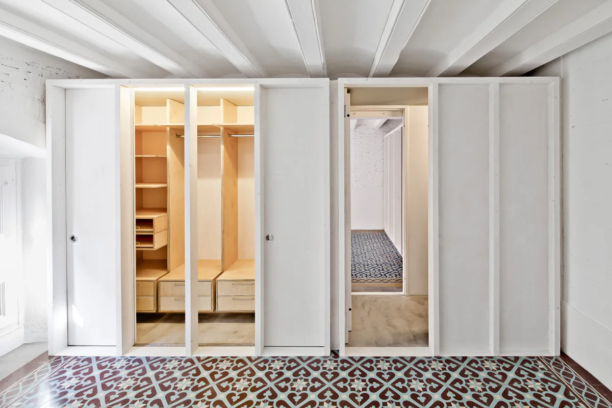 A built-in white closet with sliding doors reveals light wood shelving and drawers over a colorful patterned tile floor.