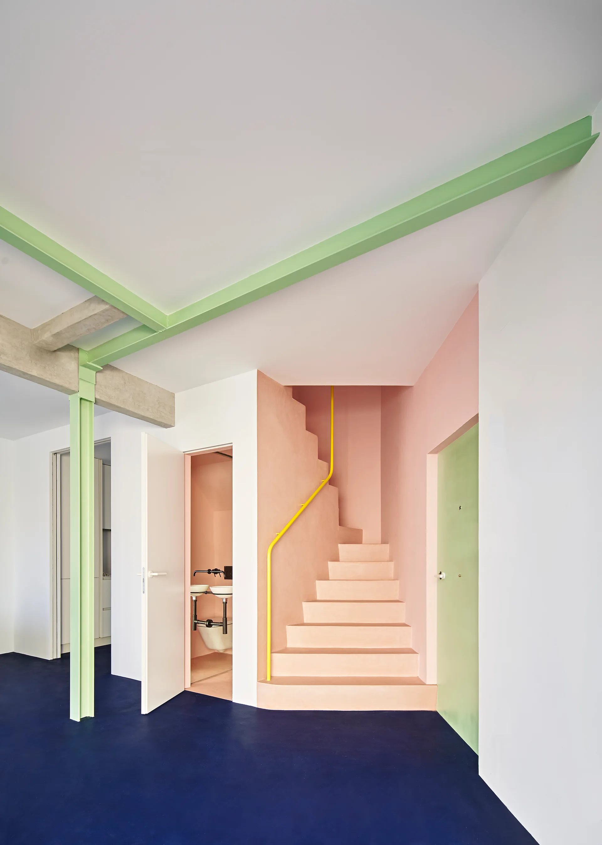 A pink concrete staircase with a yellow handrail stands beside a green structural column and beam over dark blue flooring in a white modern room.