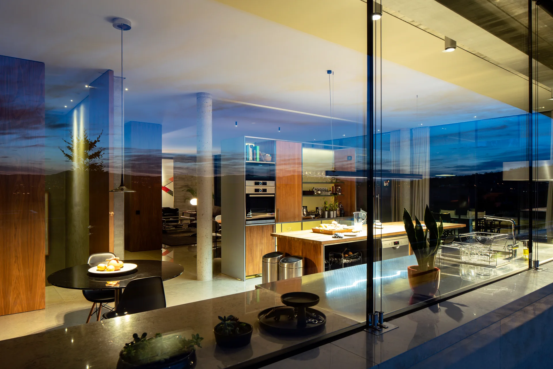 A modern kitchen with wood cabinetry, a central island, and a black dining table is viewed through a large glass wall at dusk.