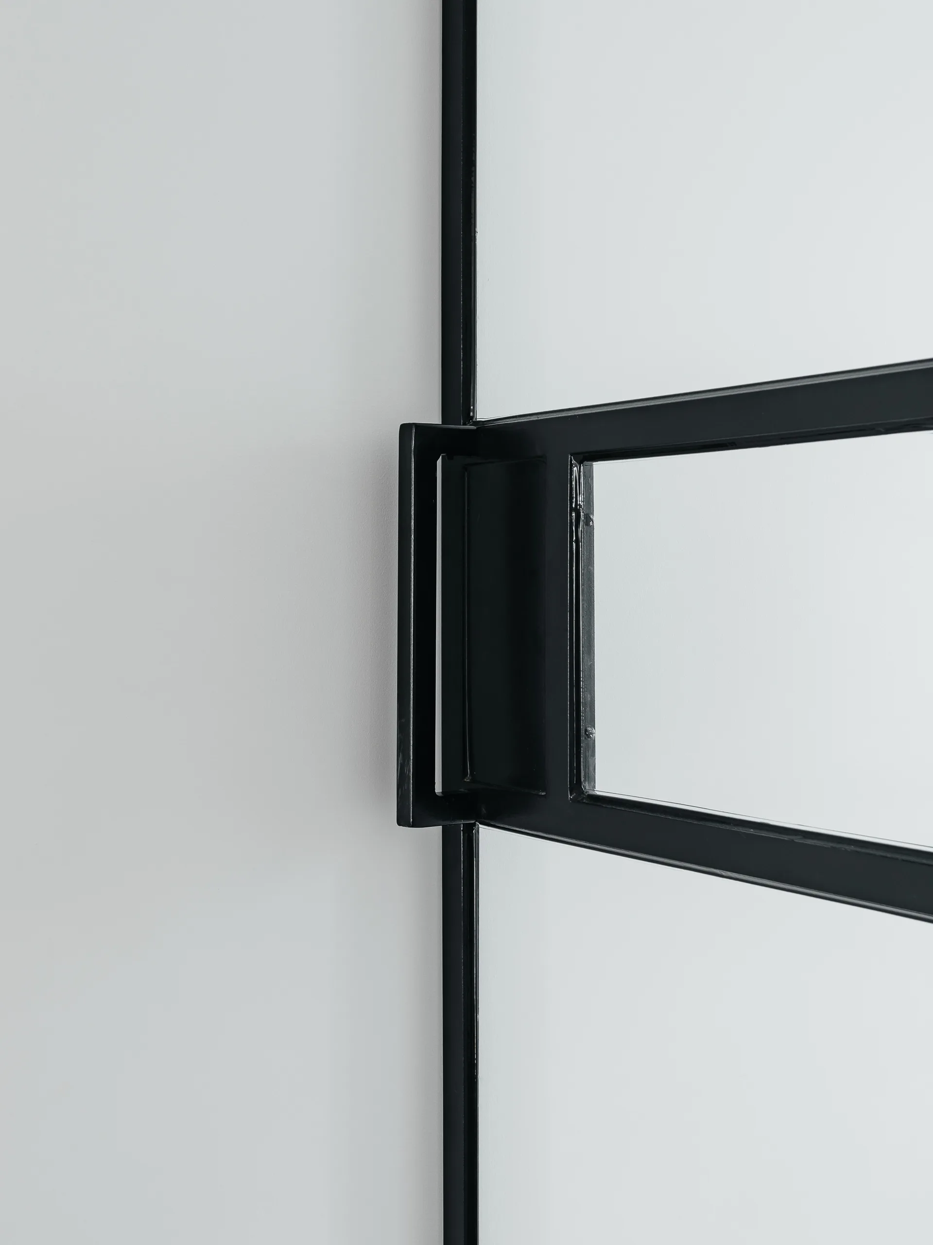 A close-up shot of a matte black metal handle mounted on a black metal door frame with a glass panel against a smooth white wall.