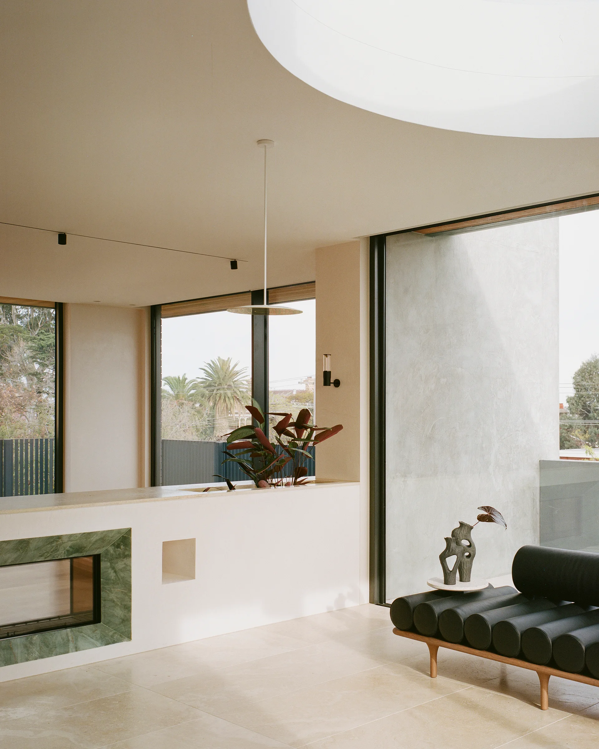 A minimalist living room featuring a green marble fireplace surround, a black cylindrical cushion daybed, and a round skylight overhead.