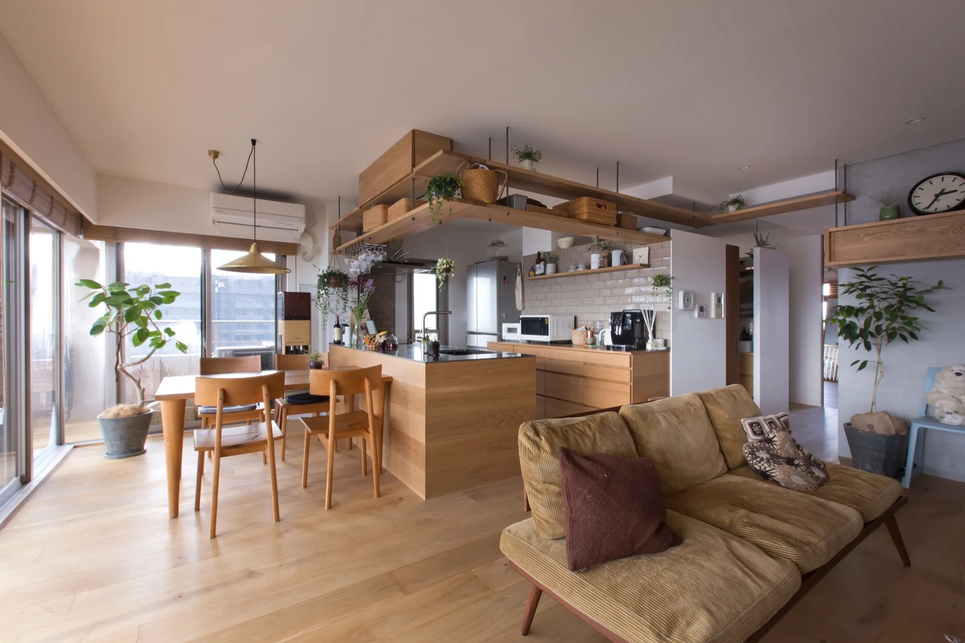 An open concept kitchen features a light wood island and dining set, a brick tile backsplash, and a corduroy sofa in the foreground.