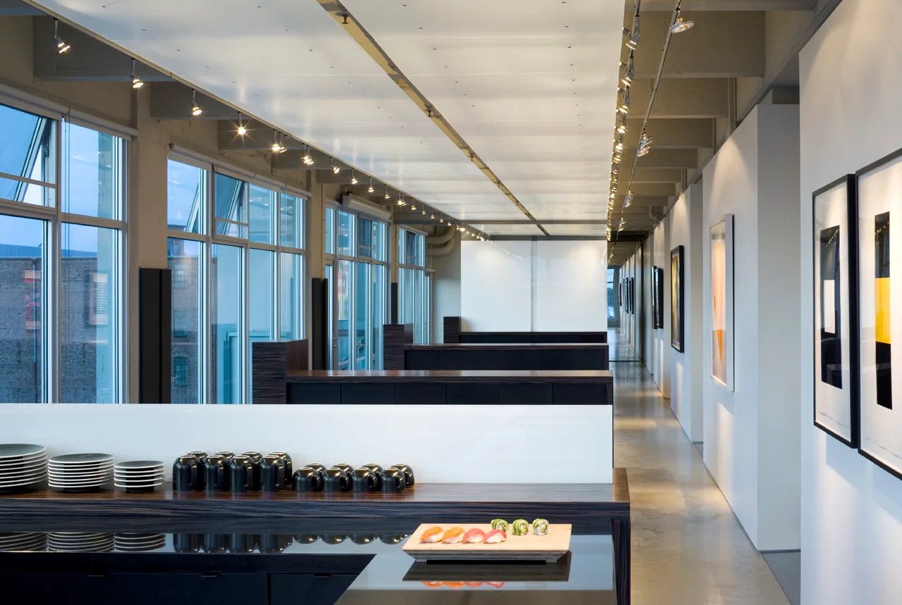 A long modern gallery corridor features large windows on one side, dark wood serving counters with plates, and framed art on white walls.