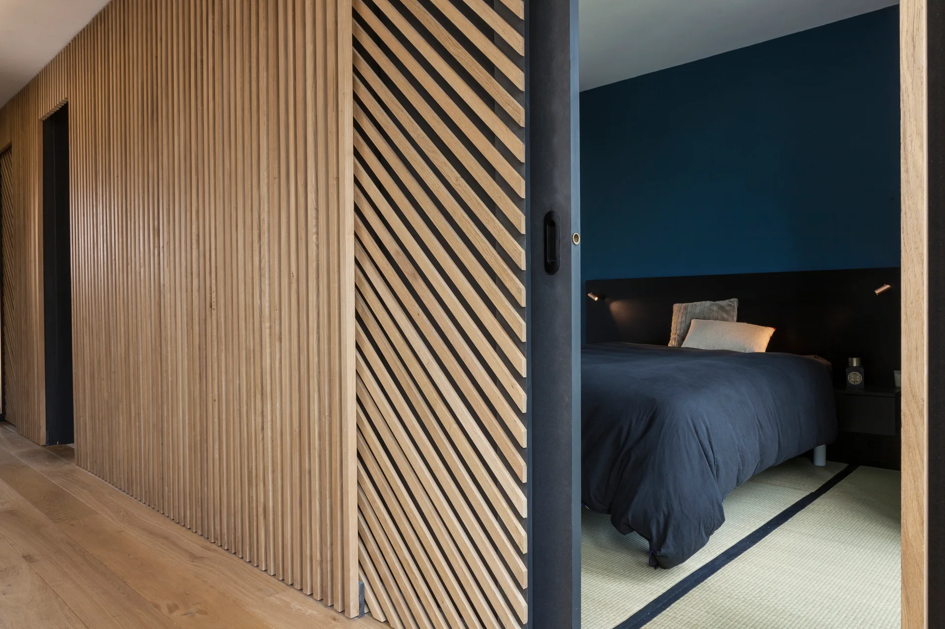 A modern Japandi bedroom with a dark blue wall and tatami mats is viewed through a sliding door featuring vertical and diagonal wood slats.