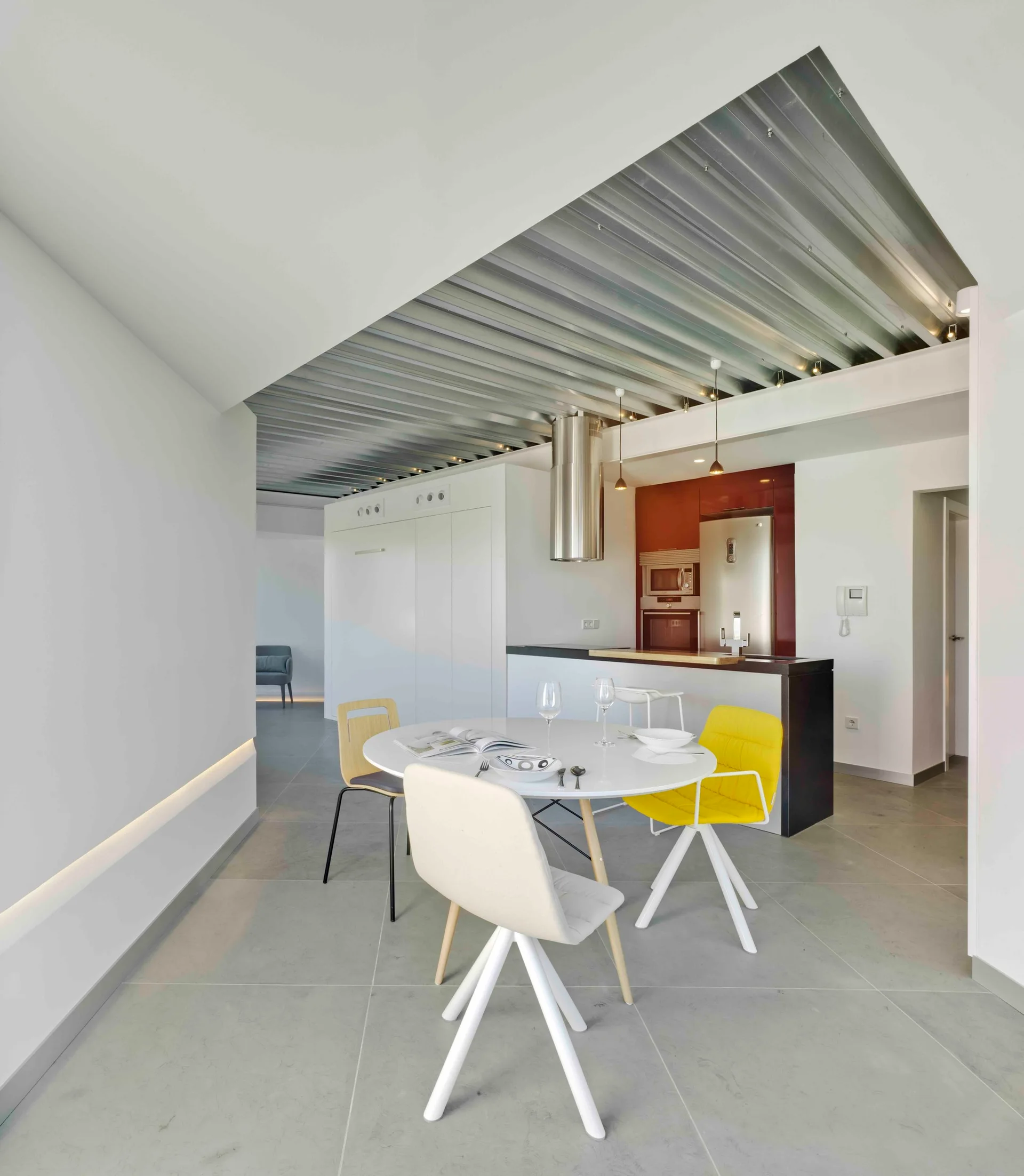A modern open-plan kitchen and dining area features a white round table, mixed chairs, and an industrial corrugated metal ceiling.