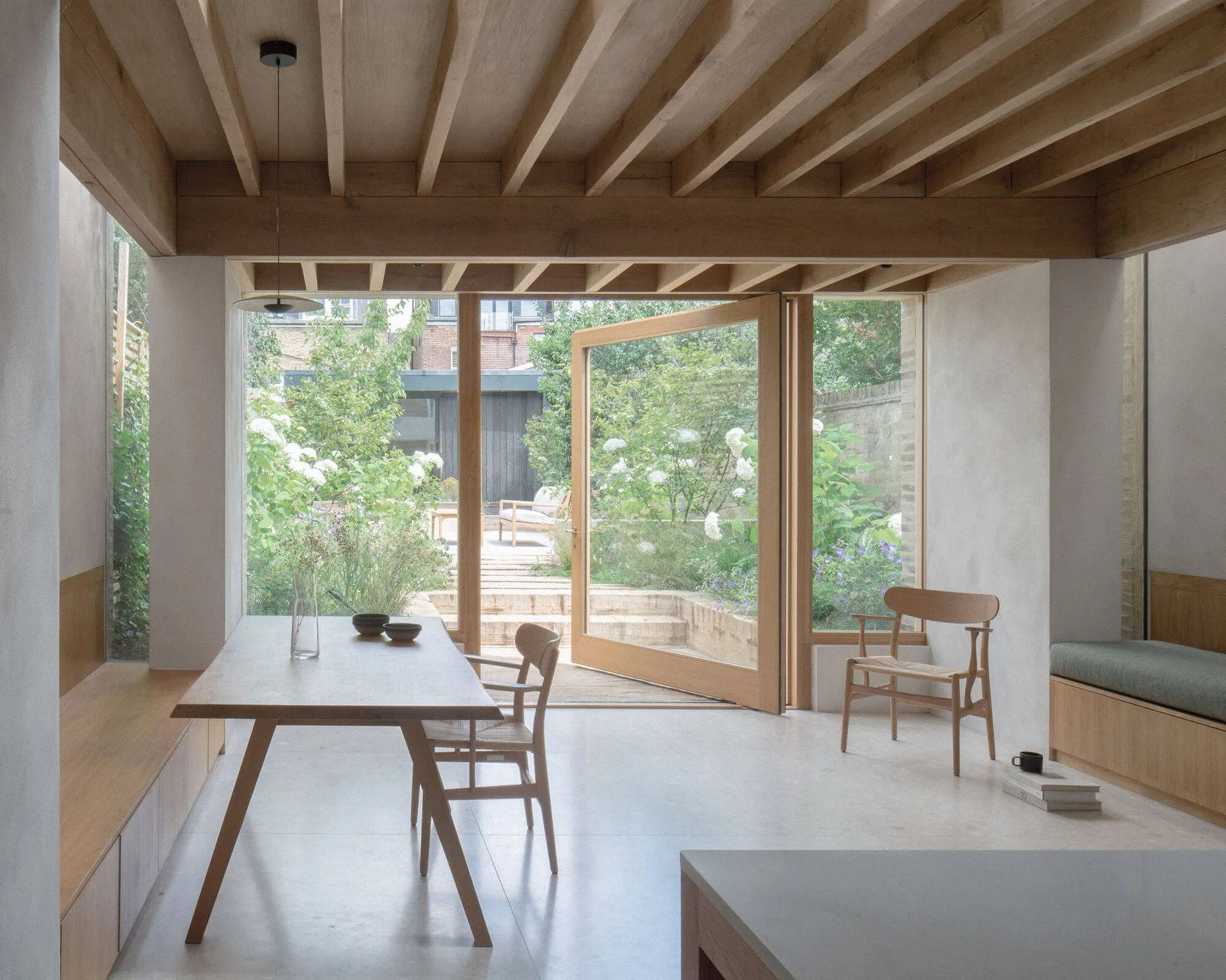 A minimalist dining room features a wooden table, exposed timber ceiling joists, and a large glass door opening to a garden.