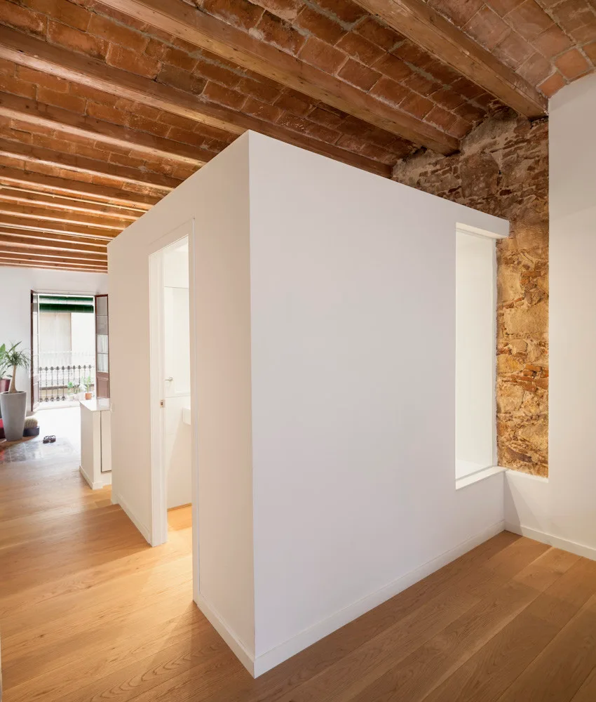 A minimalist white room partition housing a bathroom stands in a loft with exposed wood ceiling beams, hardwood floors, and a brick wall.