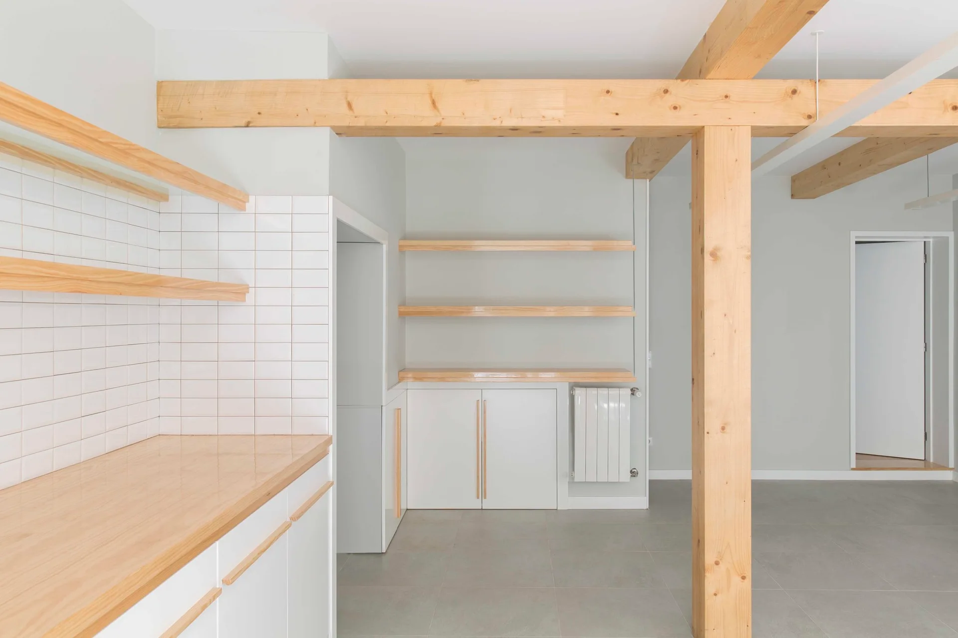 A minimalist kitchen featuring white cabinets with wood countertops, open shelving, a white tile backsplash, and exposed structural wood beams.