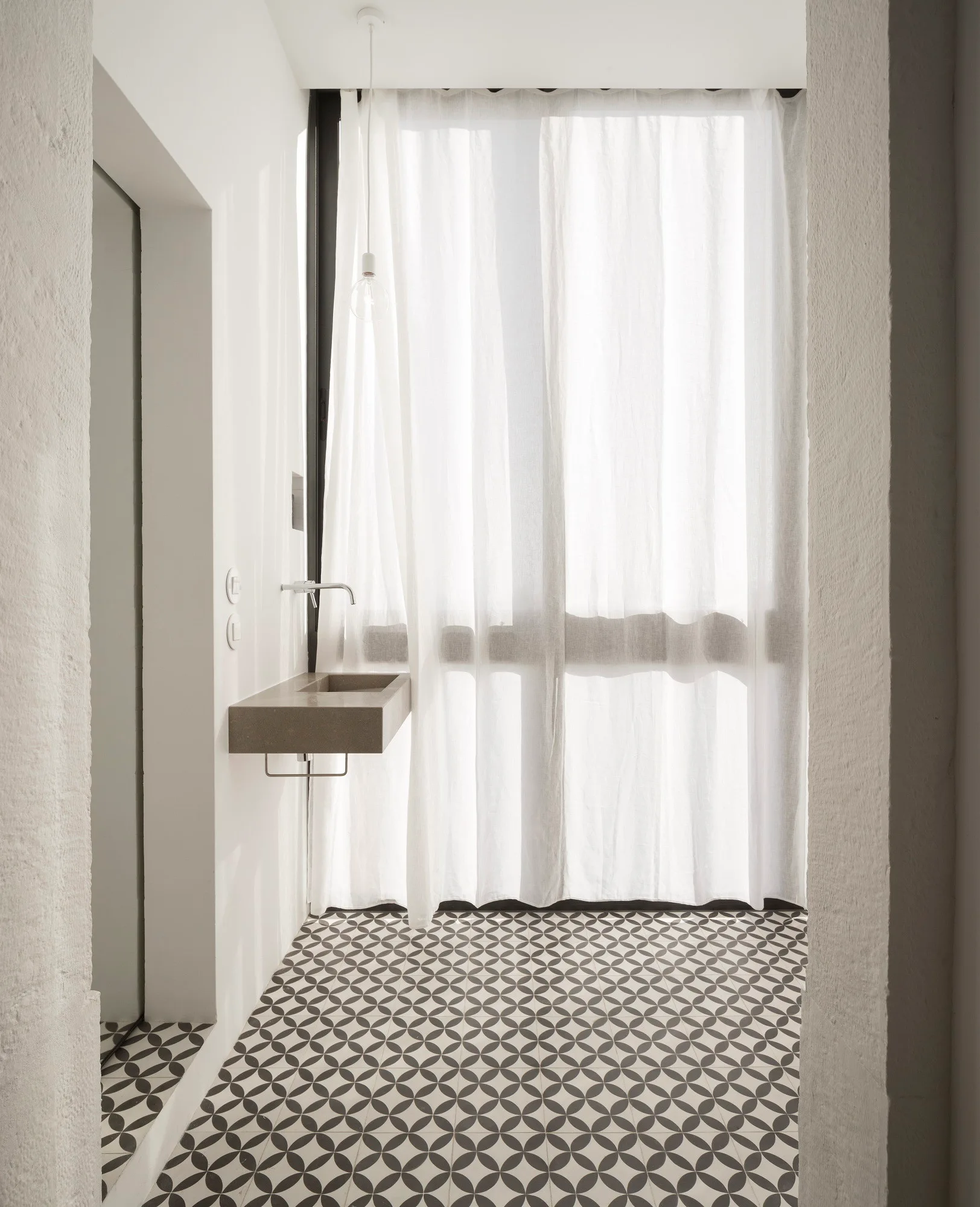 A minimalist washroom featuring a wall-mounted concrete sink, black and white patterned floor tiles, and a sheer white curtain.