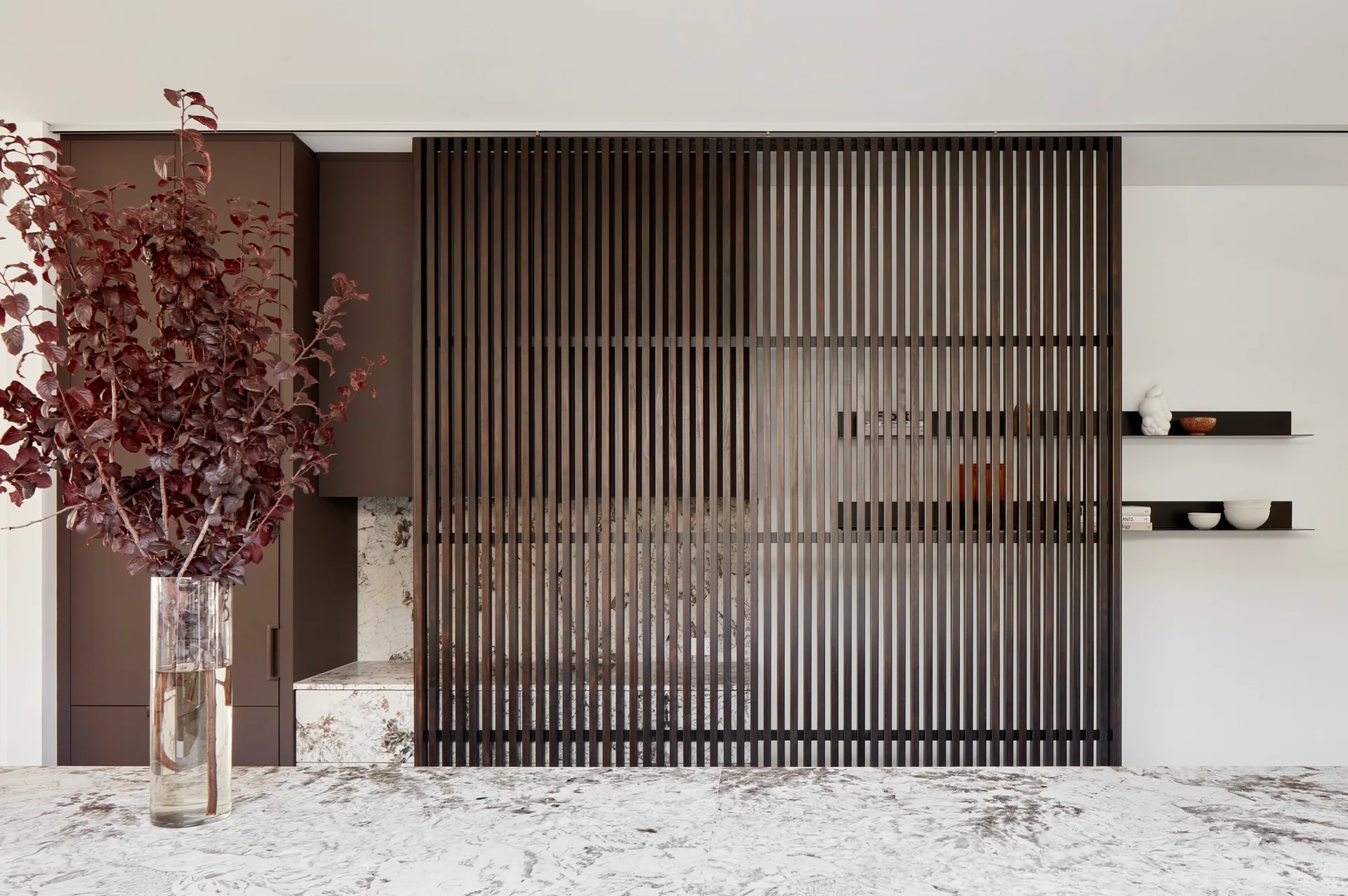 A dark wood slatted sliding screen covers a heavily veined white marble backsplash and countertop in a minimalist modern kitchen.