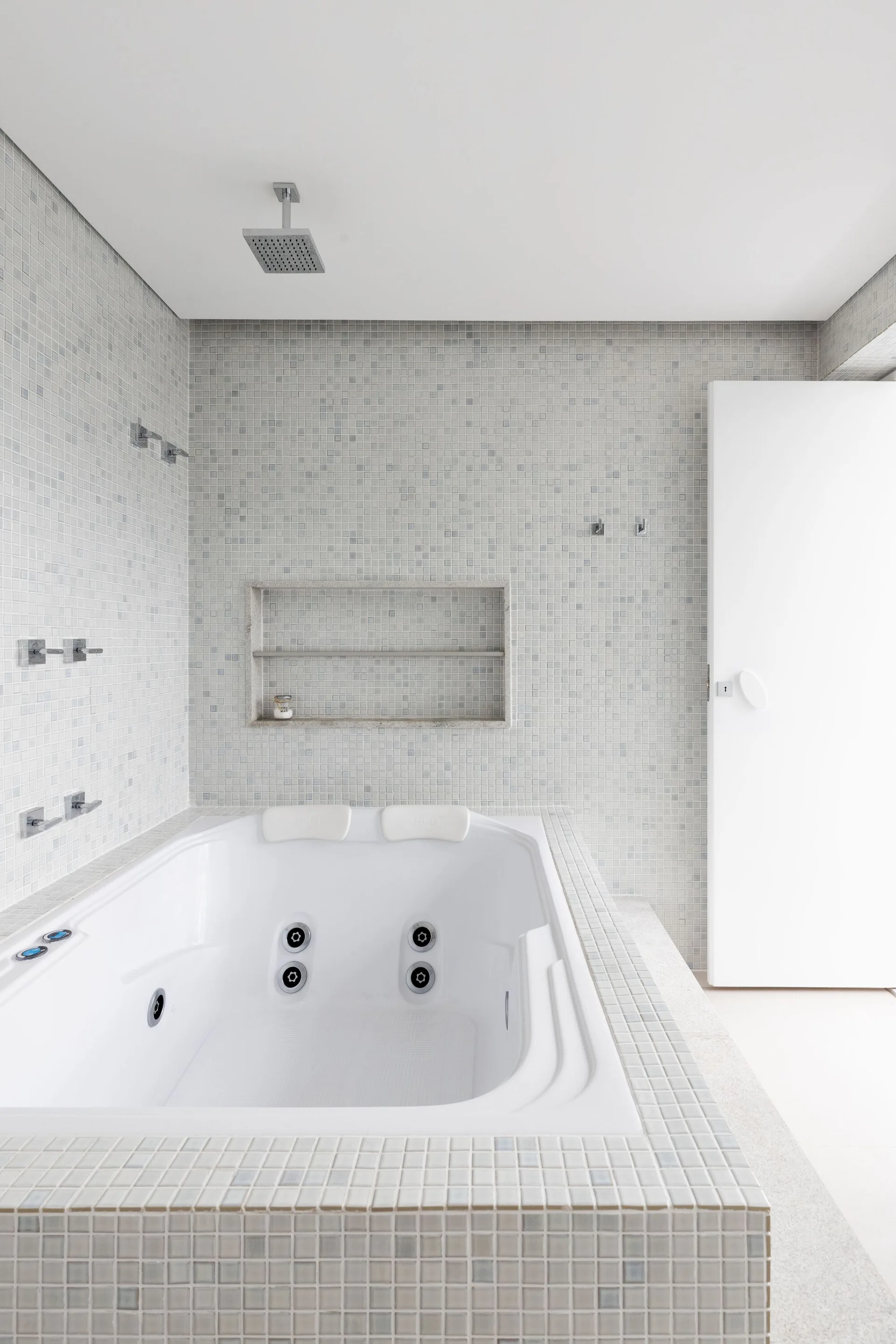 A white whirlpool bathtub is surrounded by light grey mosaic tile walls, with a recessed wall niche and a ceiling-mounted shower head.
