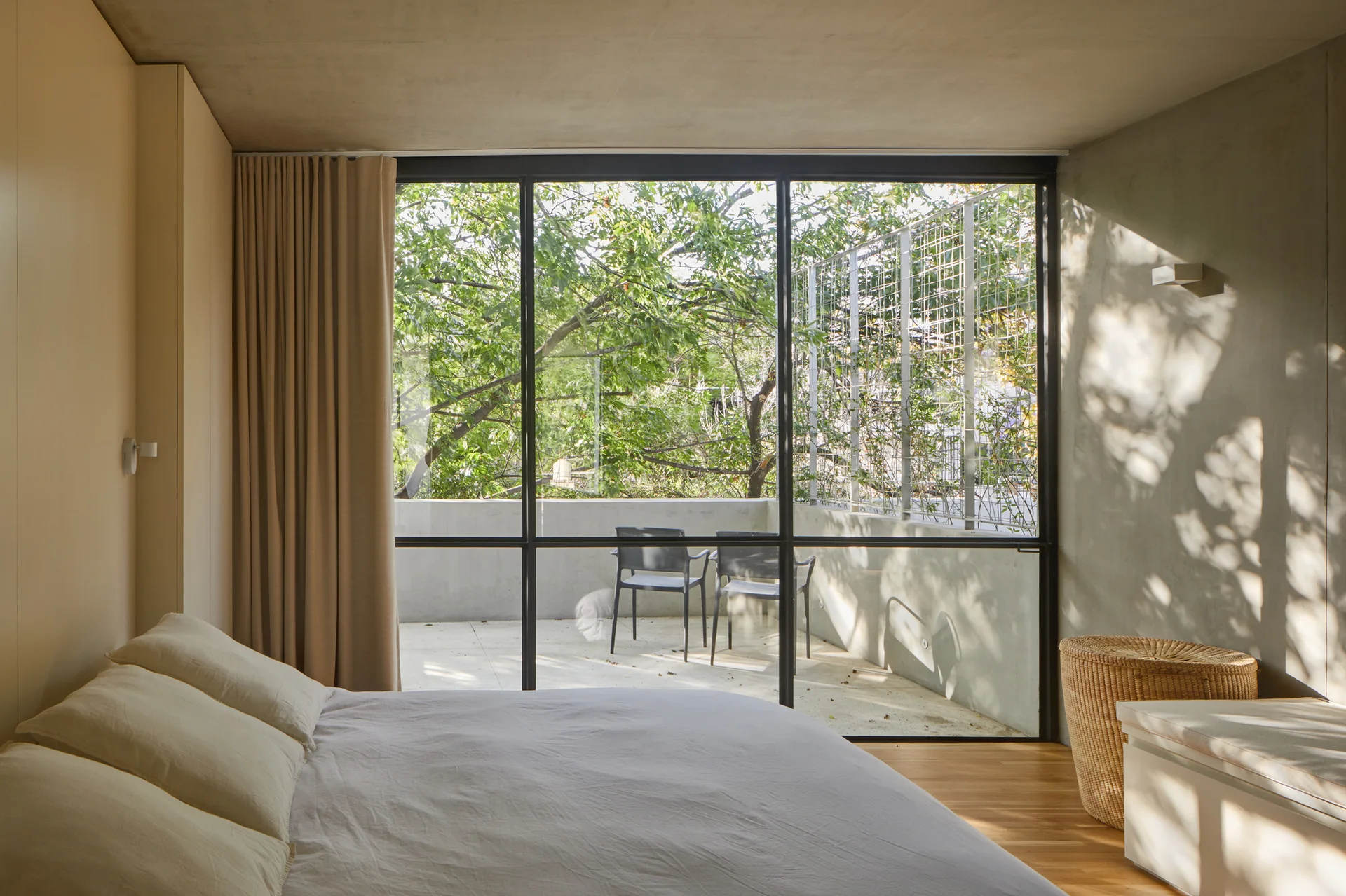 A minimalist bedroom features warm wood floors, neutral bedding, and a large metal-framed window overlooking a concrete balcony with green trees.