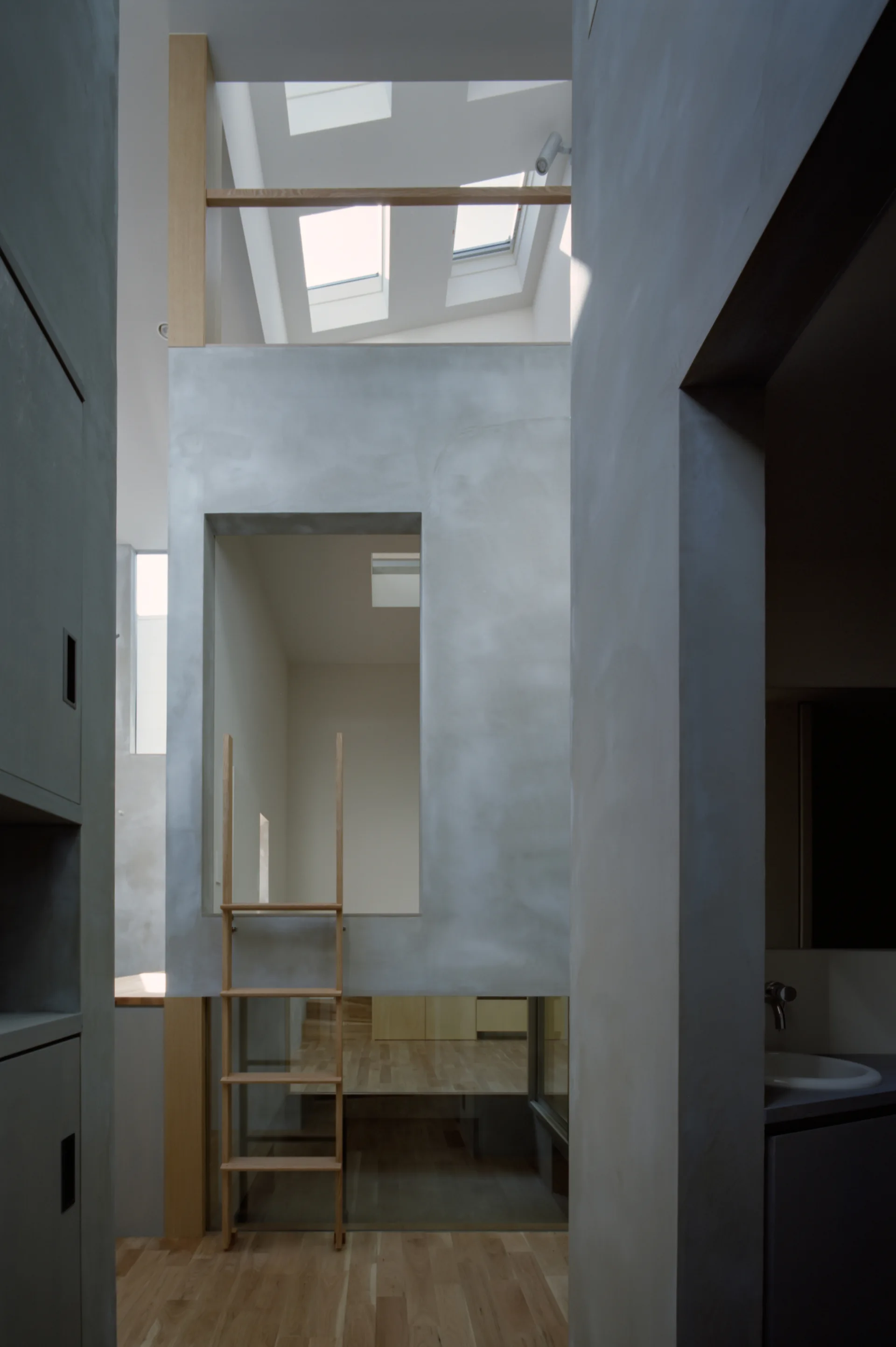 A minimalist multi-level interior featuring smooth grey plaster walls, overhead skylights, and a wooden ladder leaning against an opening.