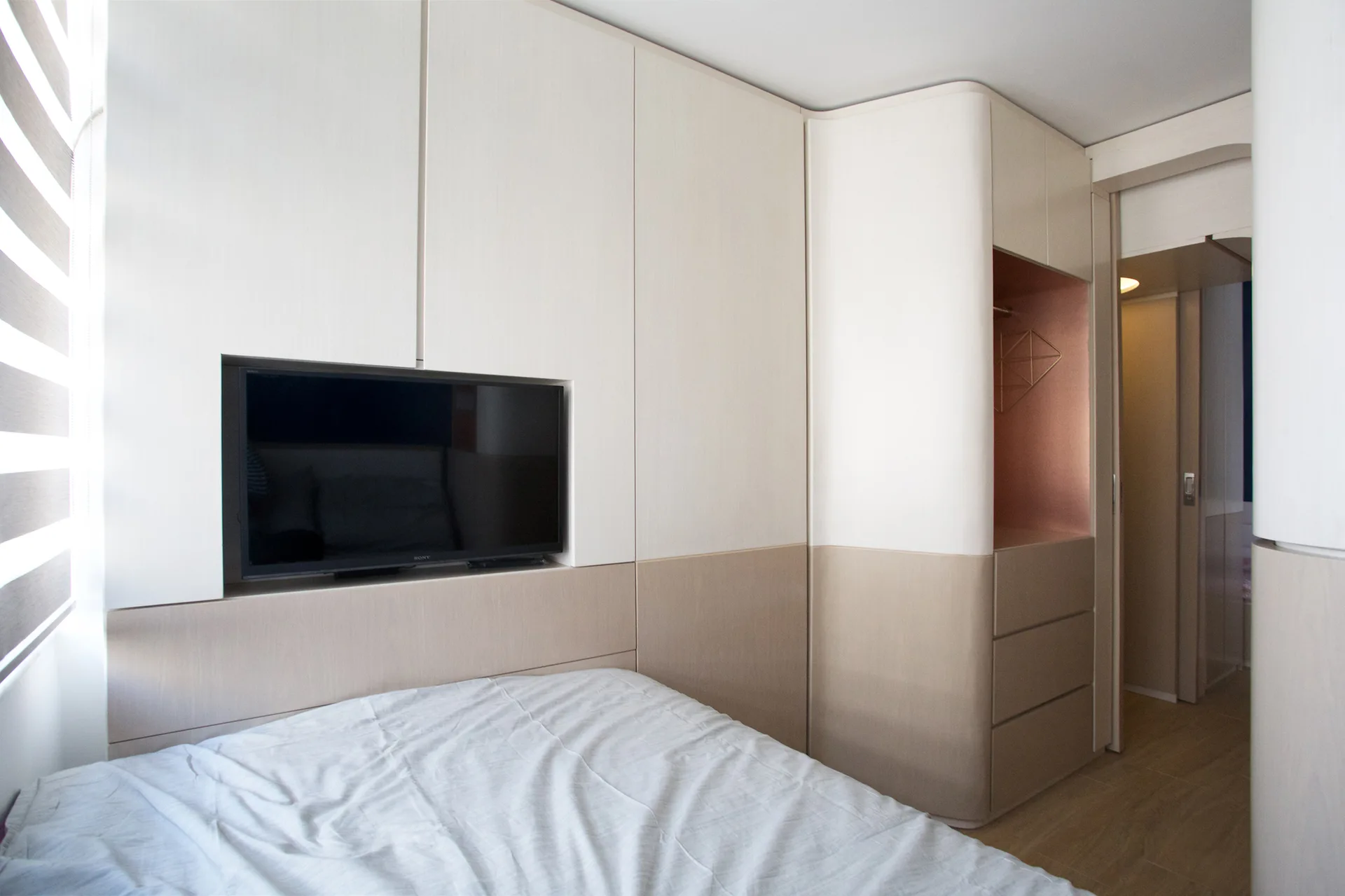 A compact bedroom features off-white custom cabinetry built around a wall-mounted television, a curved corner wardrobe, and light-colored bedding.