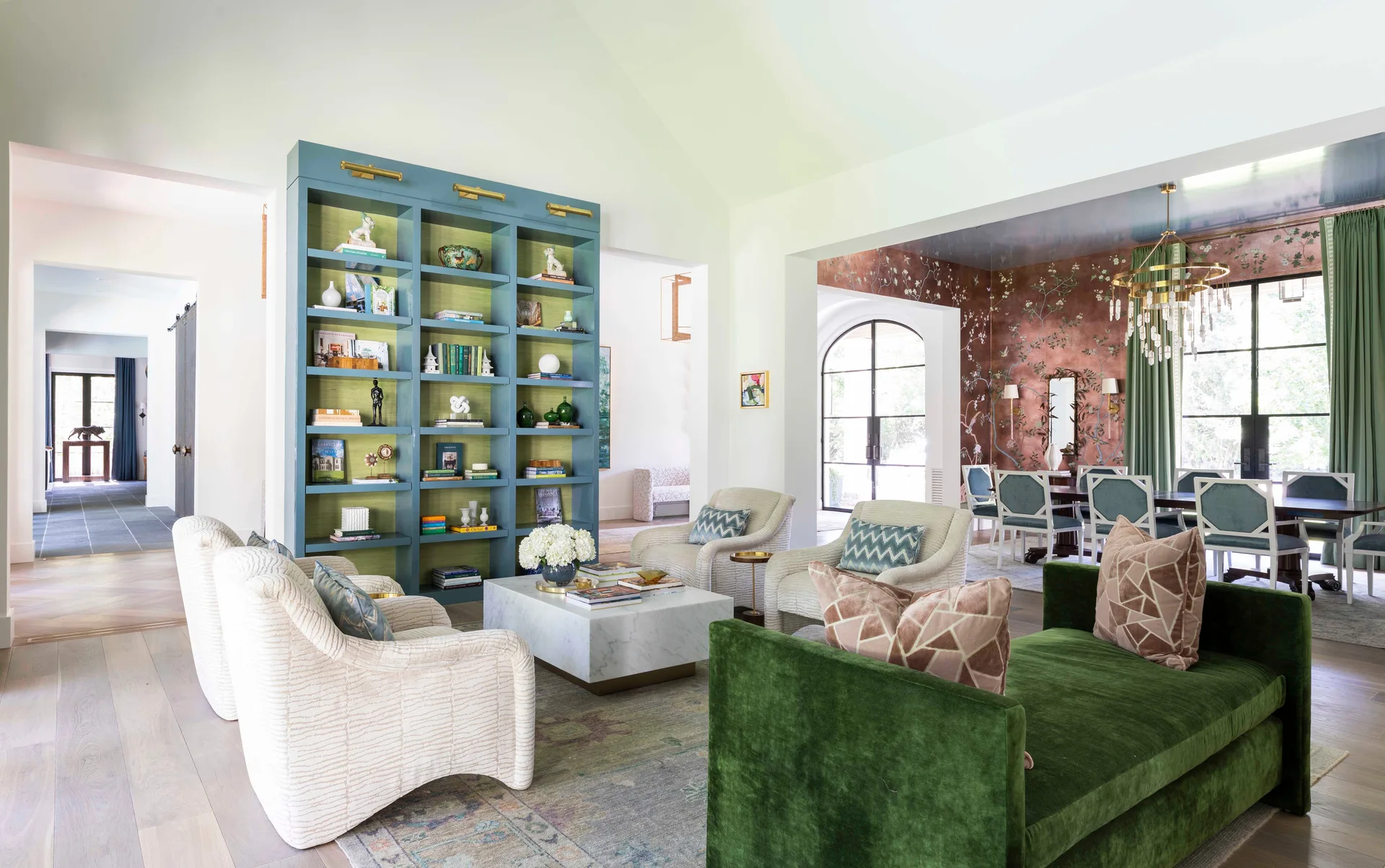A bright living room with a green velvet sofa, neutral armchairs, a large blue built-in bookshelf, and a dining area with a floral mural.