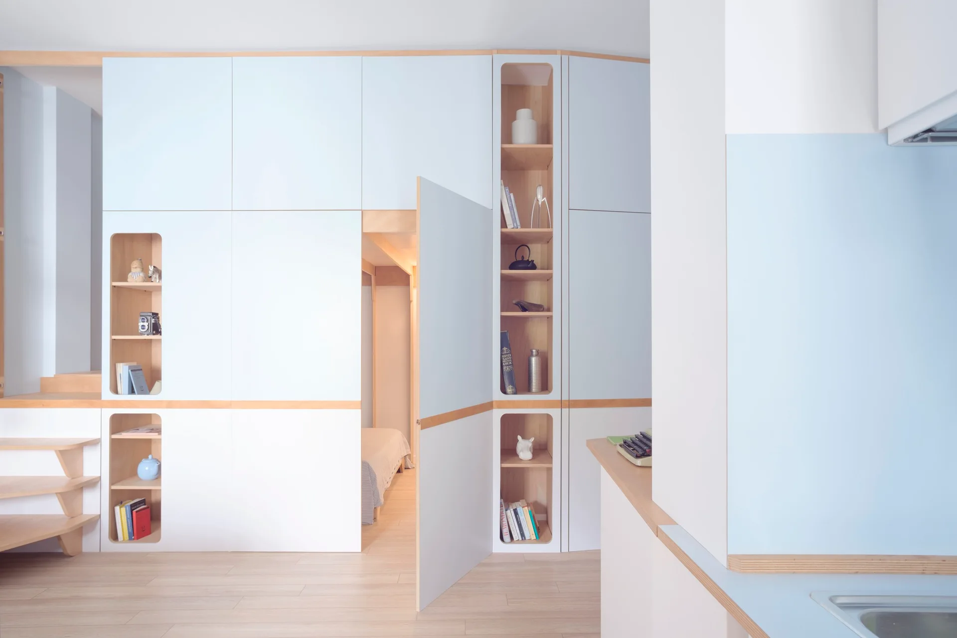 A wall of light blue and pale wood built-in cabinets features open vertical shelves and a swinging door that reveals a bedroom.