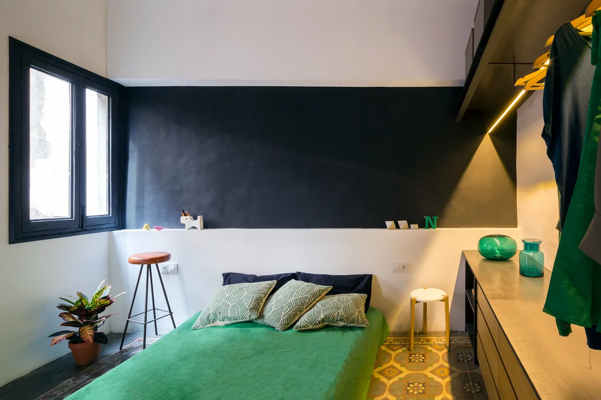 A compact bedroom featuring a green platform bed against a black and white bicolor wall, with patterned tile floors and built-in wooden storage.