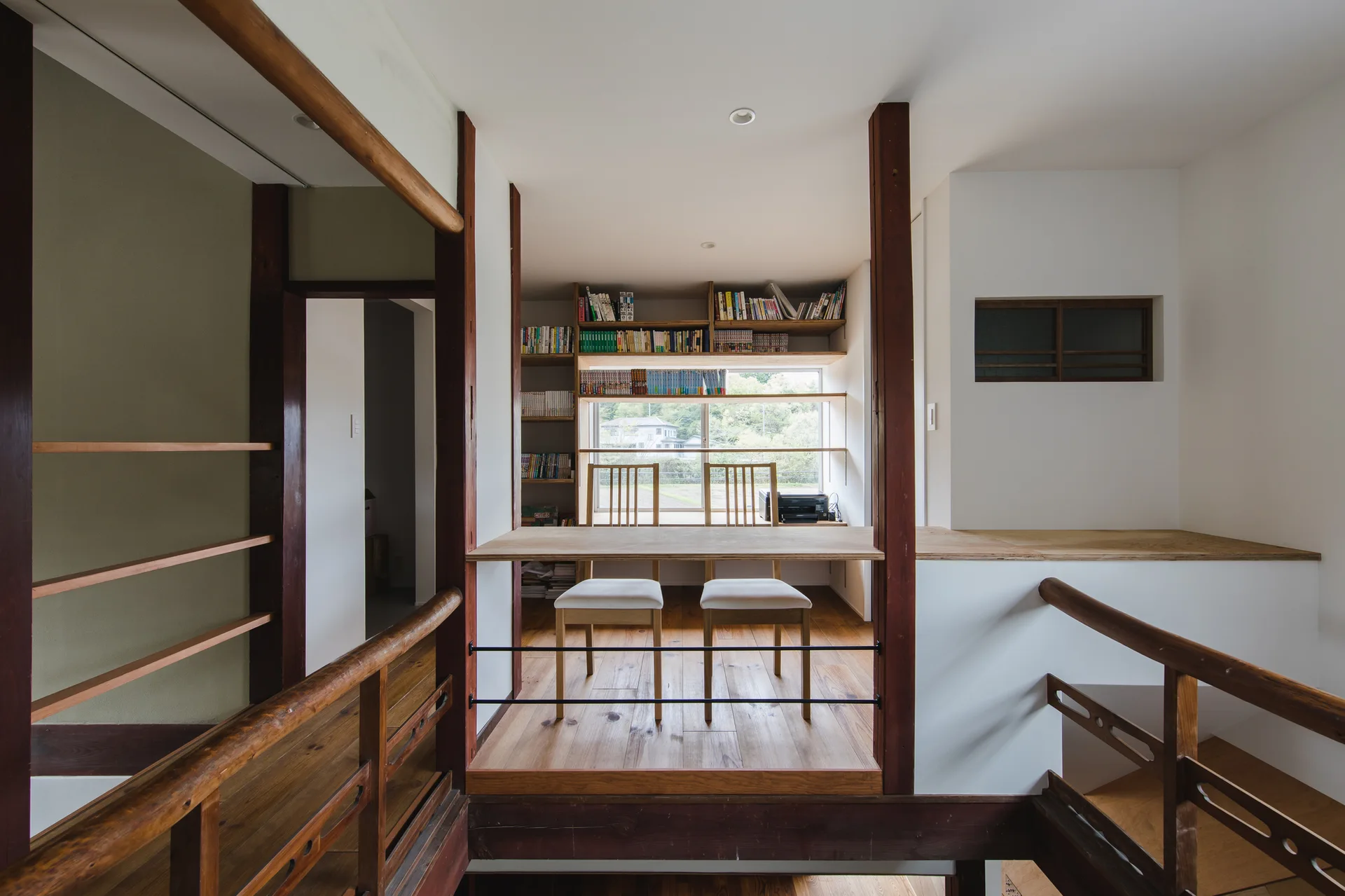 A minimalist home office features a built-in wooden desk, two chairs with light cushions, and integrated bookshelves surrounding a window.