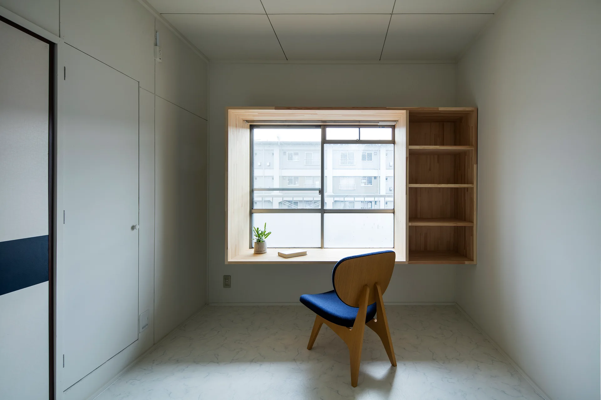 A minimalist white room features a built-in light wood window desk and shelving unit paired with a blue cushioned chair.