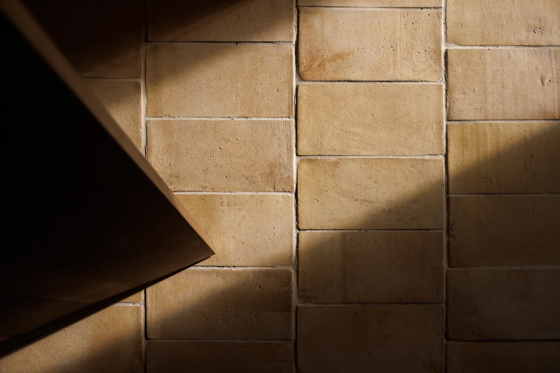 A close-up view of a matte ochre masonry block wall with horizontal grout lines and a sharp diagonal shadow cast across the textured stone surface.
