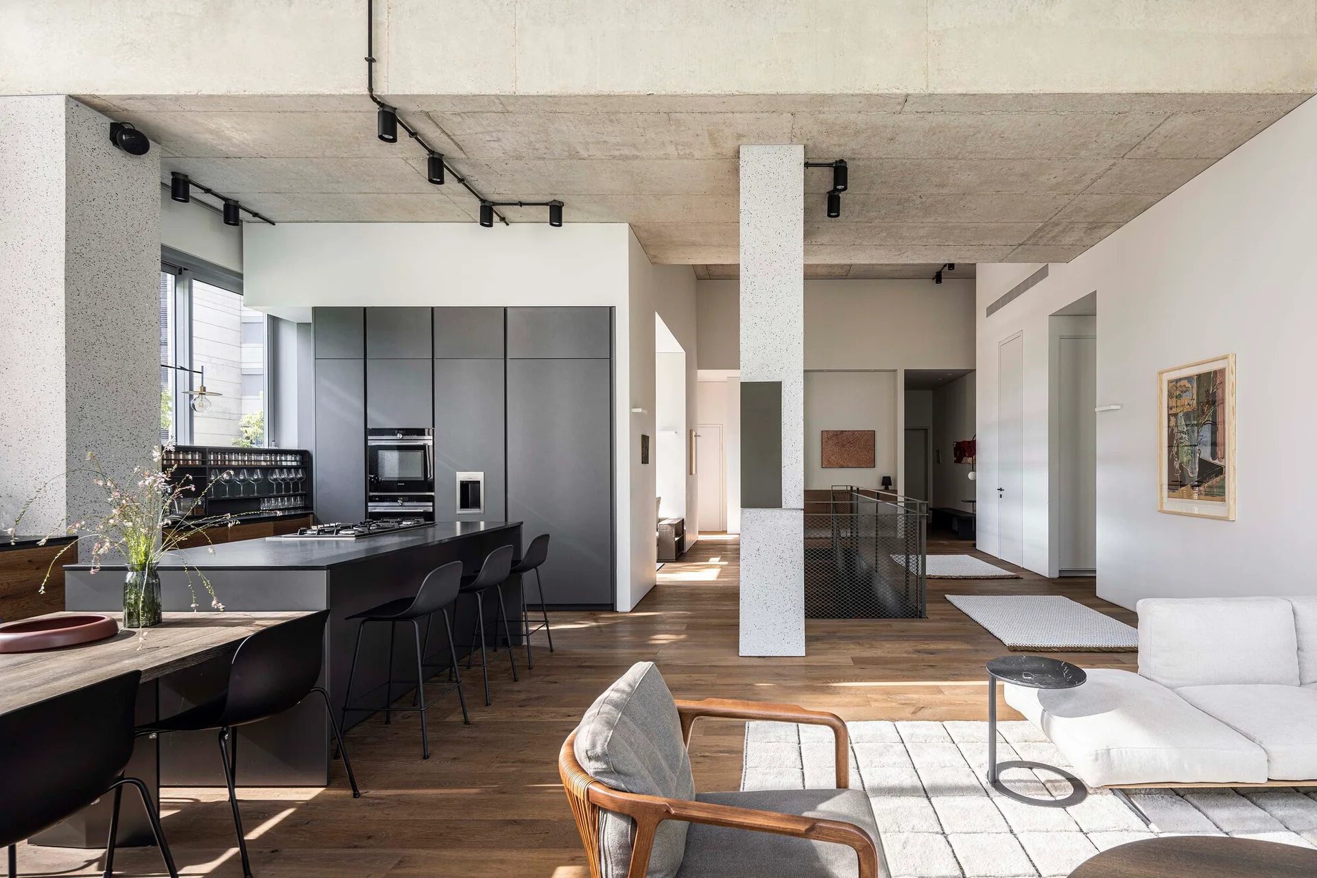 An open-concept living area features a matte grey kitchen island, an exposed concrete ceiling, a terrazzo-style support pillar, and warm wood flooring.
