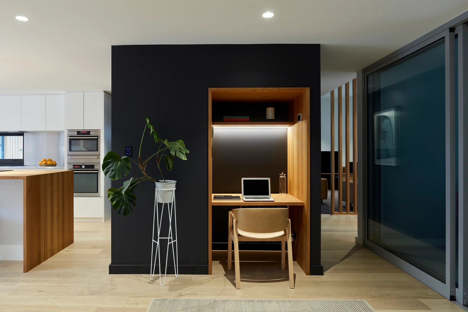 A light wood desk nook is built into a matte black feature wall next to a tall white planter, with a modern kitchen visible in the background.