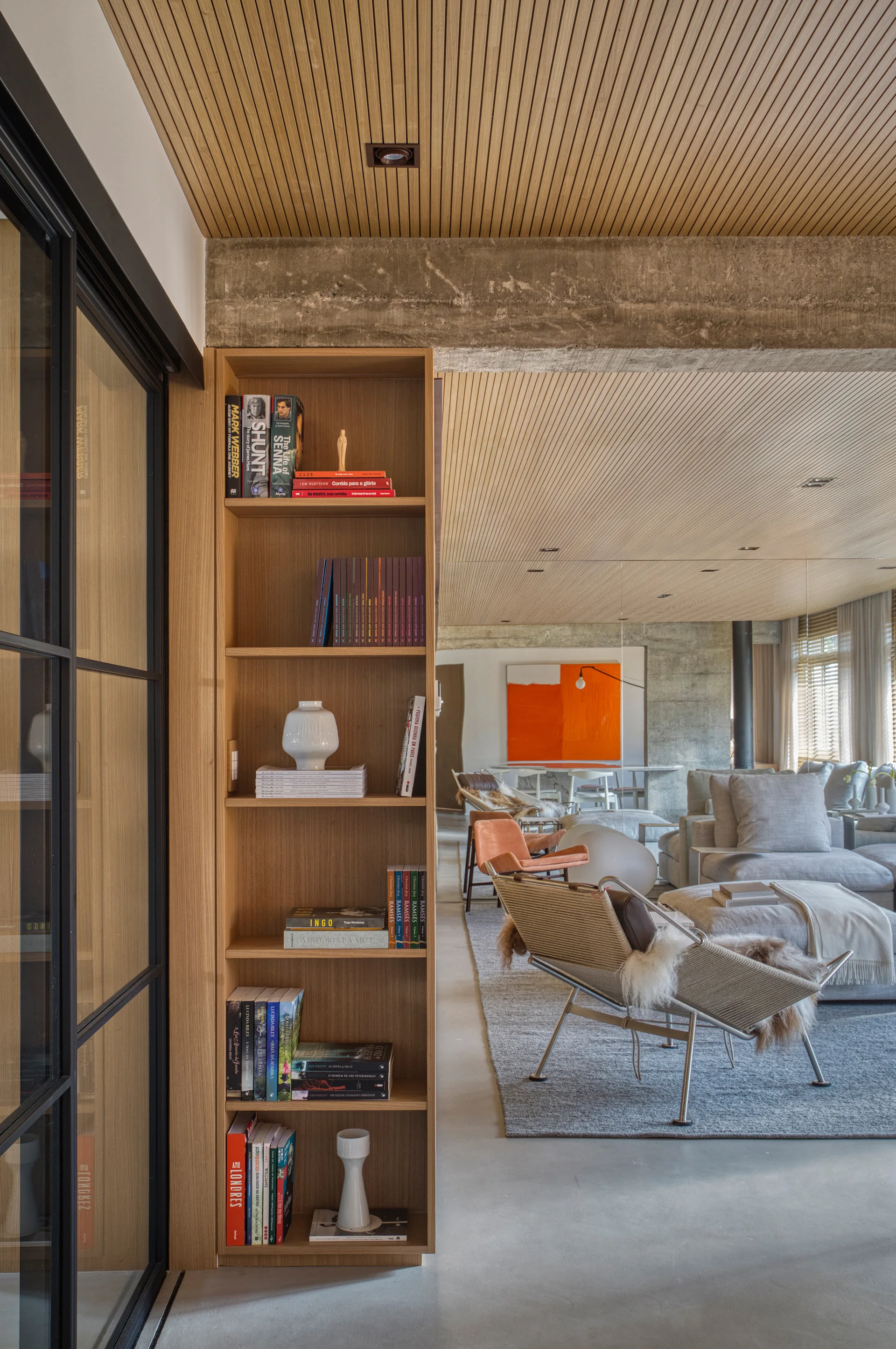 A modern living room features a tall wooden bookshelf, a slatted wood ceiling with a concrete beam, and a metal-frame lounge chair on a grey rug.